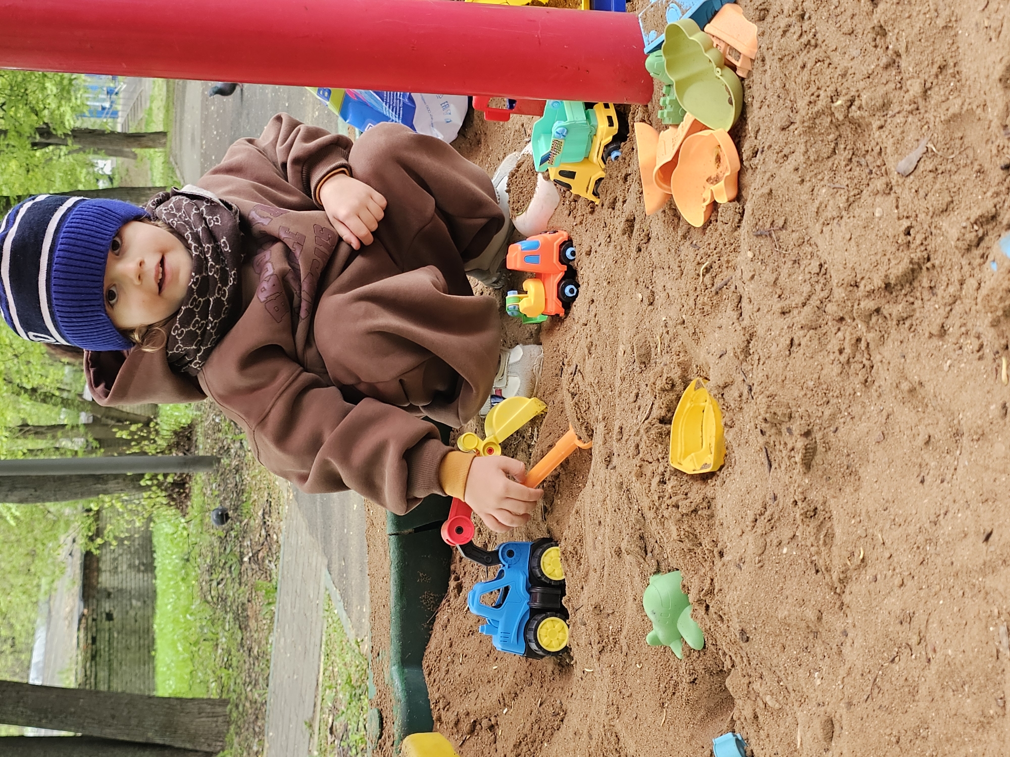 A young child wearing a blue and white striped hat, brown hoodie, and a brown Gucci scarf sits in a sandbox surrounded by colorful plastic toys. The child holds a yellow shovel and is digging in the sand. The sandbox is located in a park in Khimki, Russia, with a red metal structure visible in the background, along with trees and a paved walkway. The sandbox contains various plastic toys including trucks, shovels, and molds.