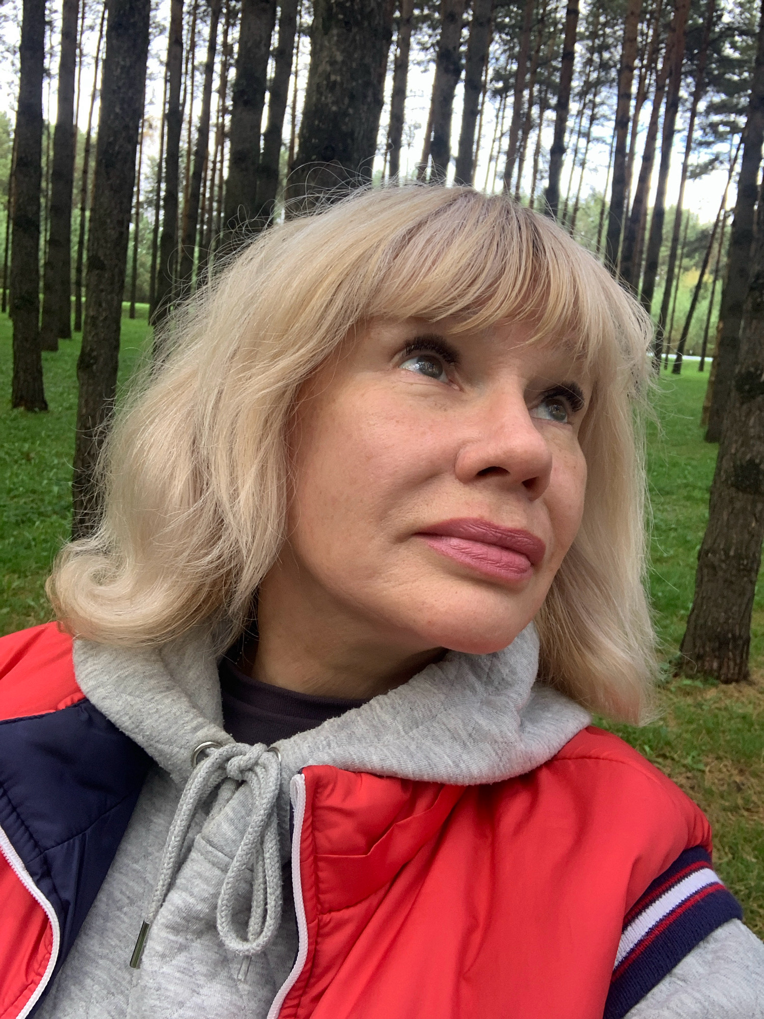 A woman with blonde hair and makeup is pictured in an outdoor setting. She is wearing a red jacket over a grey hooded sweatshirt and a dark top. The background consists of a grove of tall trees with green grass on the ground. There is no indication of the city of Prokopyevsk, Russia, within the image or its surrounding context.