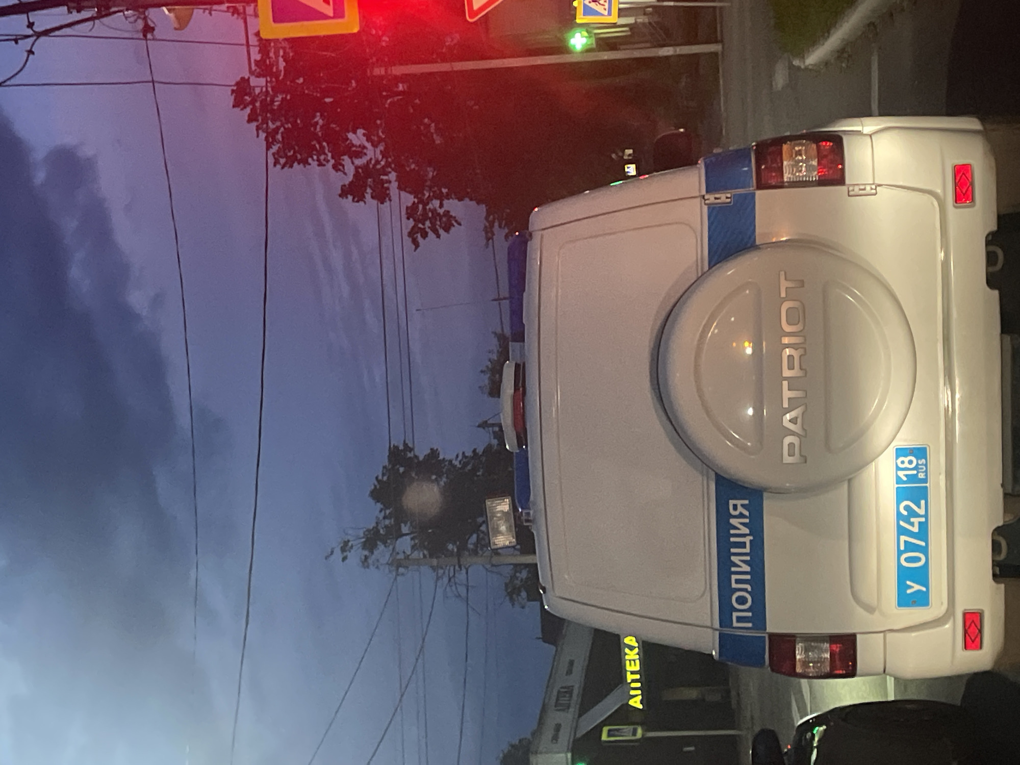 This image shows the rear of a white police vehicle, likely a UAZ Patriot, parked on a street at dusk or night. The vehicle has blue and white markings with the word "ПОЛИЦИЯ" (Police) visible in blue lettering. A spare tire is mounted on the rear door, with the word "PATRIOT" embossed on it. The license plate is blue with white characters and reads "у 0742 18 RUS." Red and blue flashing lights are visible on the roof of the vehicle.
In the background, there is a building with a sign that says "АПTEKA" (Apotheka, meaning pharmacy). Traffic lights are visible, with one showing a red signal. Trees and power lines are also present. The overall atmosphere suggests a street scene in a Russian city, consistent with Votkinsk. There are no people clearly visible in the image.