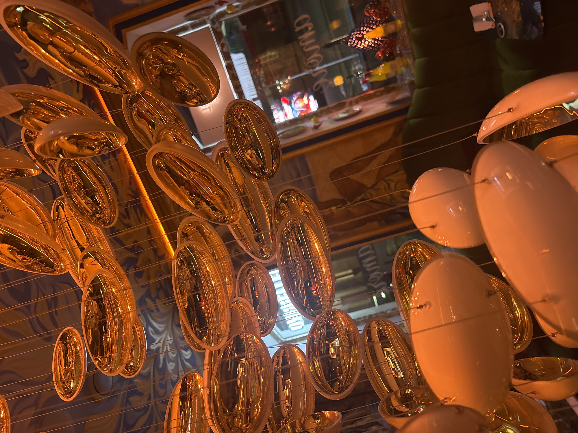This is a low-angle, close-up shot of a cluster of hanging light fixtures, likely in an indoor setting. The lights are a mix of ovoid and oval shapes, with some featuring a reflective, metallic gold finish and others a matte white ceramic. They are suspended by thin wires from an unseen ceiling, which is decorated with a swirling blue and white pattern.
In the background, through a glass window or partition, the scene appears to be a restaurant or bar. A green upholstered sofa is visible, as are bottles on a shelf, and reflections in the glass suggest the presence of other patrons or streetlights outside. One of the oval lights in the foreground reflects a sign with Cyrillic script, possibly reading "ОМБРЕ", which is consistent with a location in Minsk, Belarus. The lighting in the image is warm and ambient, casting a golden glow on the surrounding objects. There are no people clearly visible or interacting in the frame.
