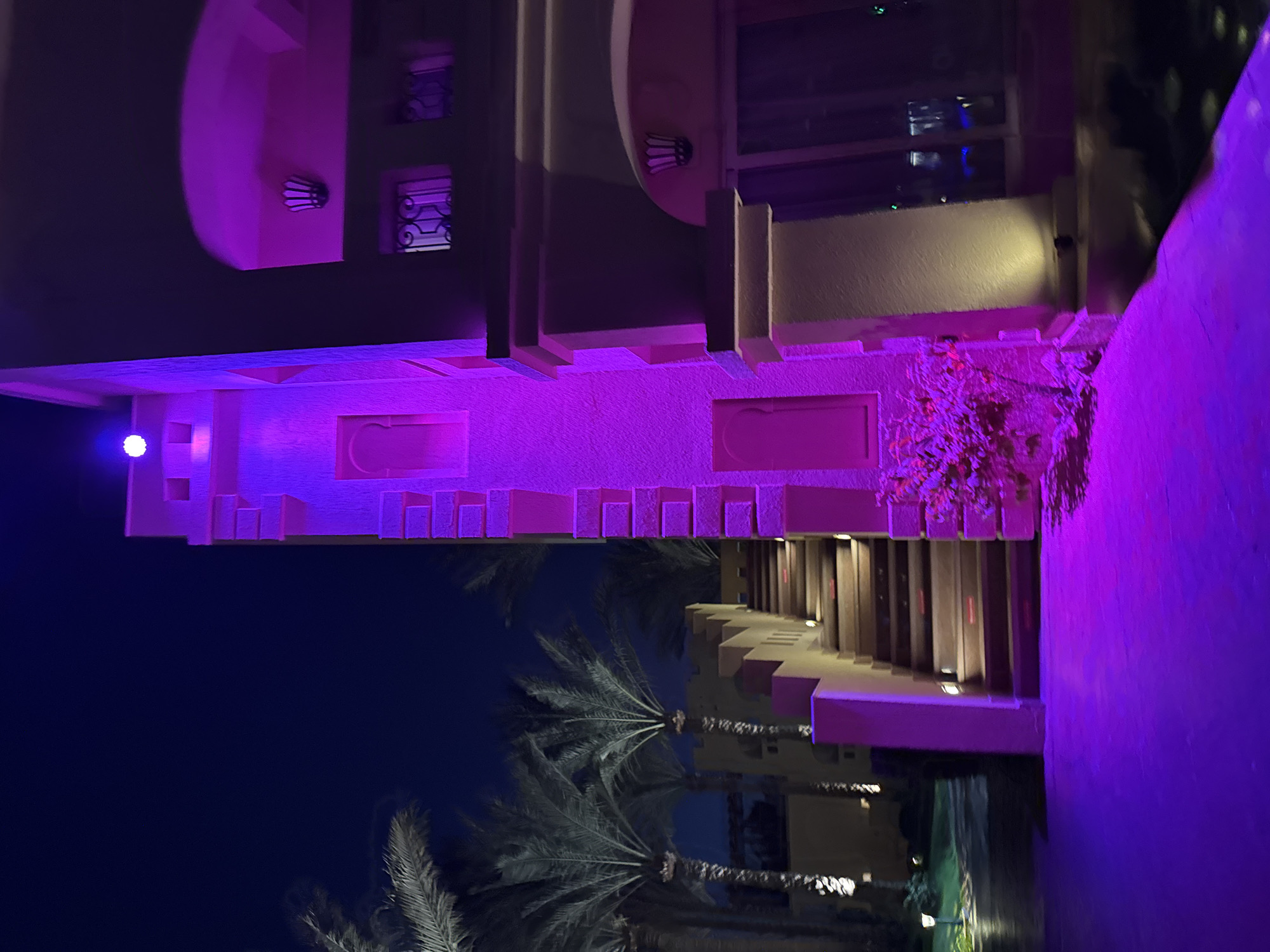The image depicts an exterior view of a building at night, illuminated by purple and yellow lights. The architecture appears to be in a Middle Eastern or North African style, with decorative elements on the facade and ornate window grills. Palm trees are visible in the background, suggesting a warm climate. A set of stairs leads up to an entrance, also lit by ambient lighting. There are no people or animals visible in the scene. The overall atmosphere is one of nighttime ambiance and possibly a resort or hotel setting. The location is specified as Unknown, Egypt.