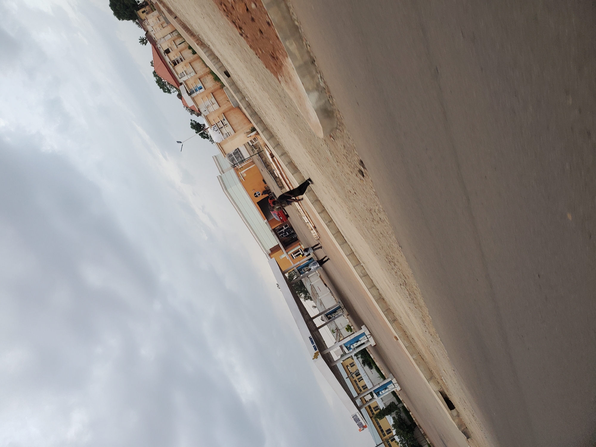 The image shows a gas station located in the city of Bukuru, Nigeria. The gas station has a white canopy over multiple fuel pumps. Two individuals are visible near the pumps, with one walking away from the camera and the other standing. In the background, there are several buildings, including a tan-colored structure with a red roof. A red car is parked near one of the buildings. The sky is overcast with gray clouds. The road in front of the gas station is paved, and there is a dirt and gravel area to the right of the road.