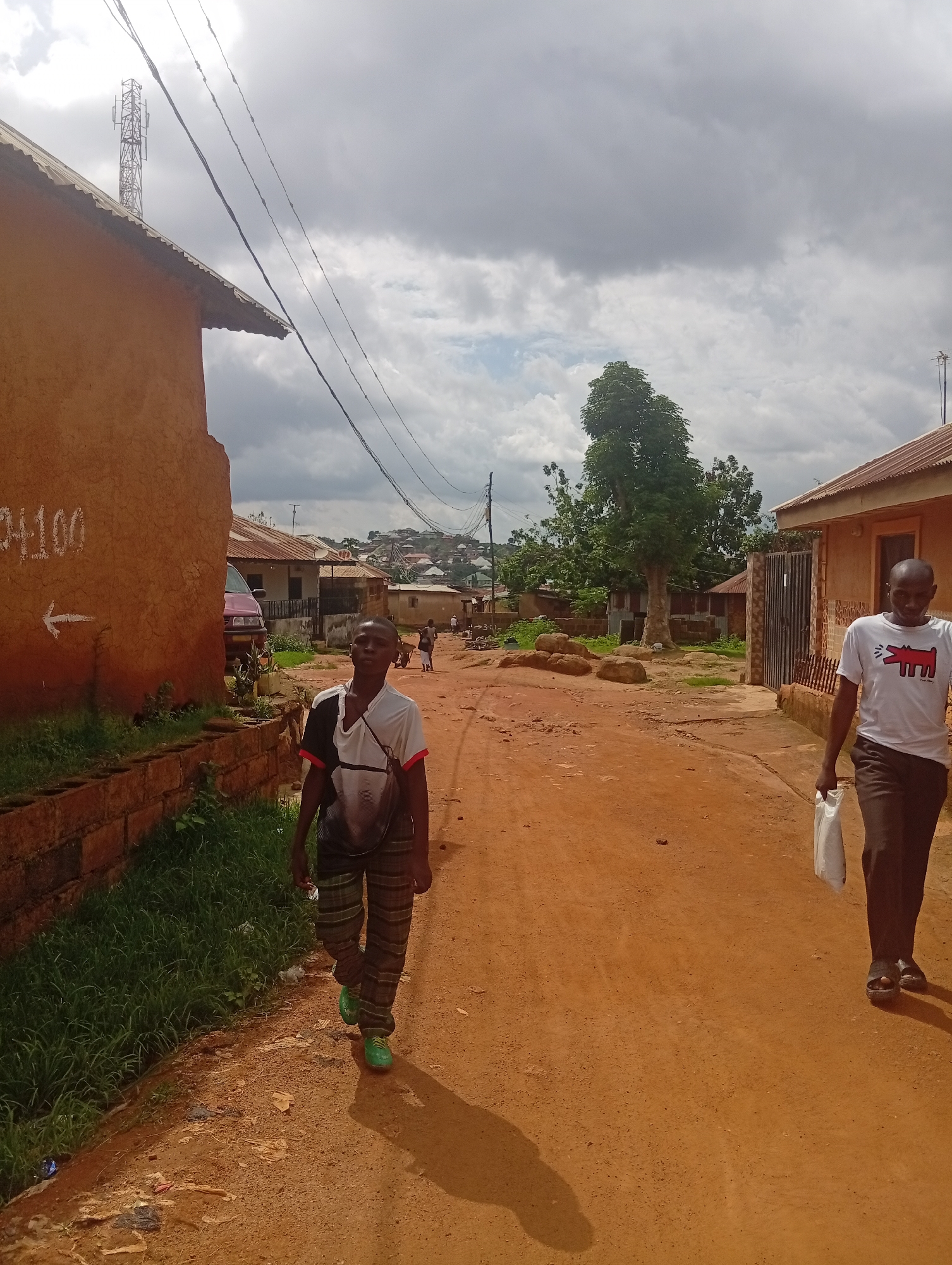 The image depicts an outdoor scene in Jos, Nigeria, on a cloudy day. Two men are walking on a dirt road. The man in the foreground, on the left side of the road, is a young boy wearing a black and white t-shirt with a patterned design on the sleeves, and checkered pants with green shoes. He is looking towards the camera. On the right side of the road, a man is walking away from the camera, carrying a white plastic bag in his right hand. He is wearing a white t-shirt with a red graphic and dark pants.
The setting appears to be a residential area with buildings constructed from earth or mud. A telecommunication tower is visible in the background, along with scattered houses and trees. The road is unpaved and reddish-brown in color. There is a wall with grass growing on top to the left of the young boy. Some debris is scattered on the side of the road. The sky is heavily clouded, suggesting potential for rain. The overall atmosphere appears casual and everyday.
White text with an arrow pointing left is visible on the orange-colored wall on the left side of the image, reading "1100".