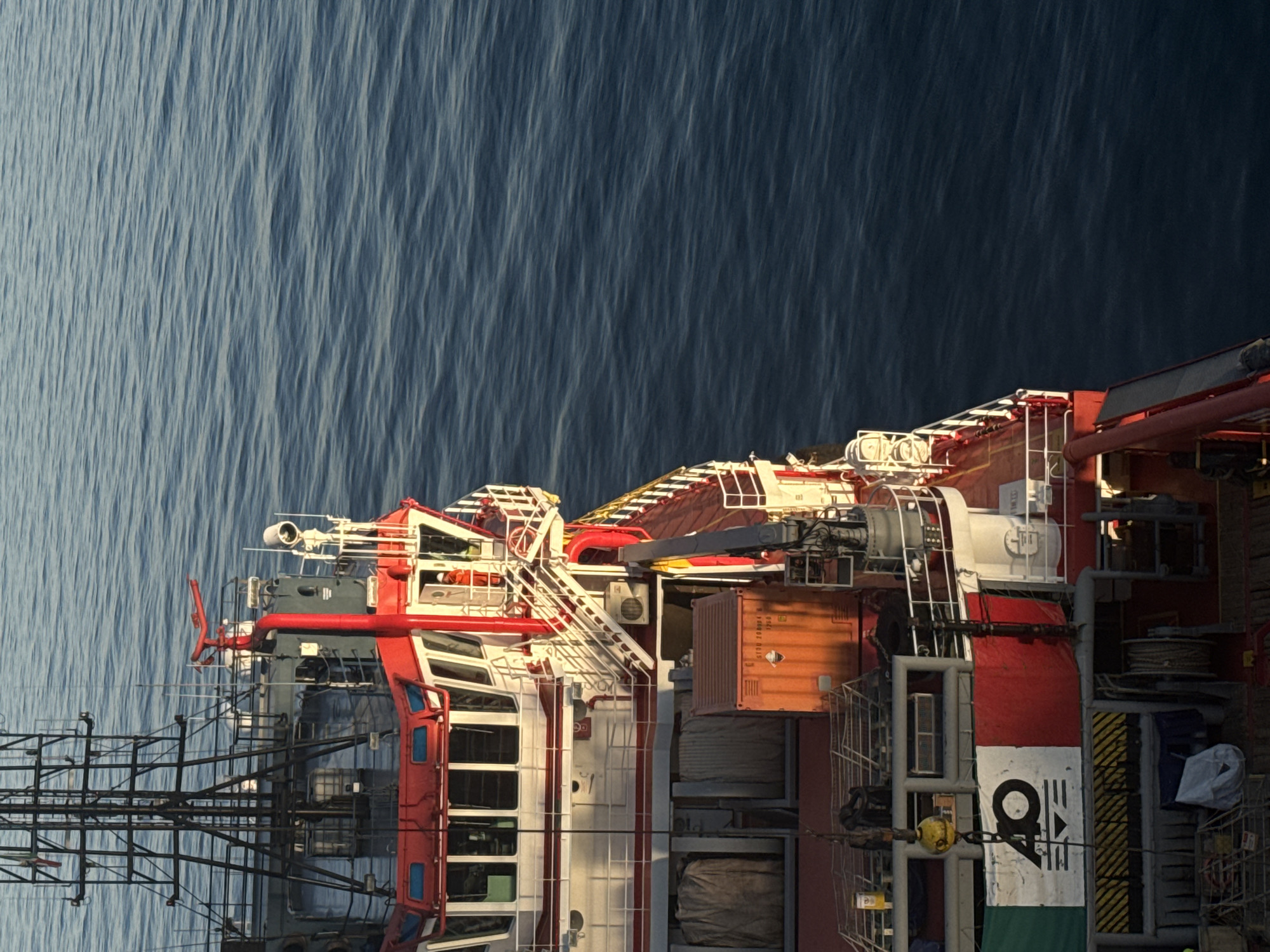 This is a high-angle, medium shot of the stern of a red and white vessel sailing on a deep blue, calm sea. The vessel has a prominent red superstructure with multiple levels, white railings, and numerous windows. A large black mast structure is visible on the upper left of the ship. A person in an orange uniform is visible on an external stairway on the right side of the superstructure. An orange shipping container is mounted on the deck. Part of the Italian flag is visible on the port side of the vessel, near the stern. The lighting suggests it is either early morning or late afternoon, with sunlight casting long shadows. The image is focused on the vessel and the water.