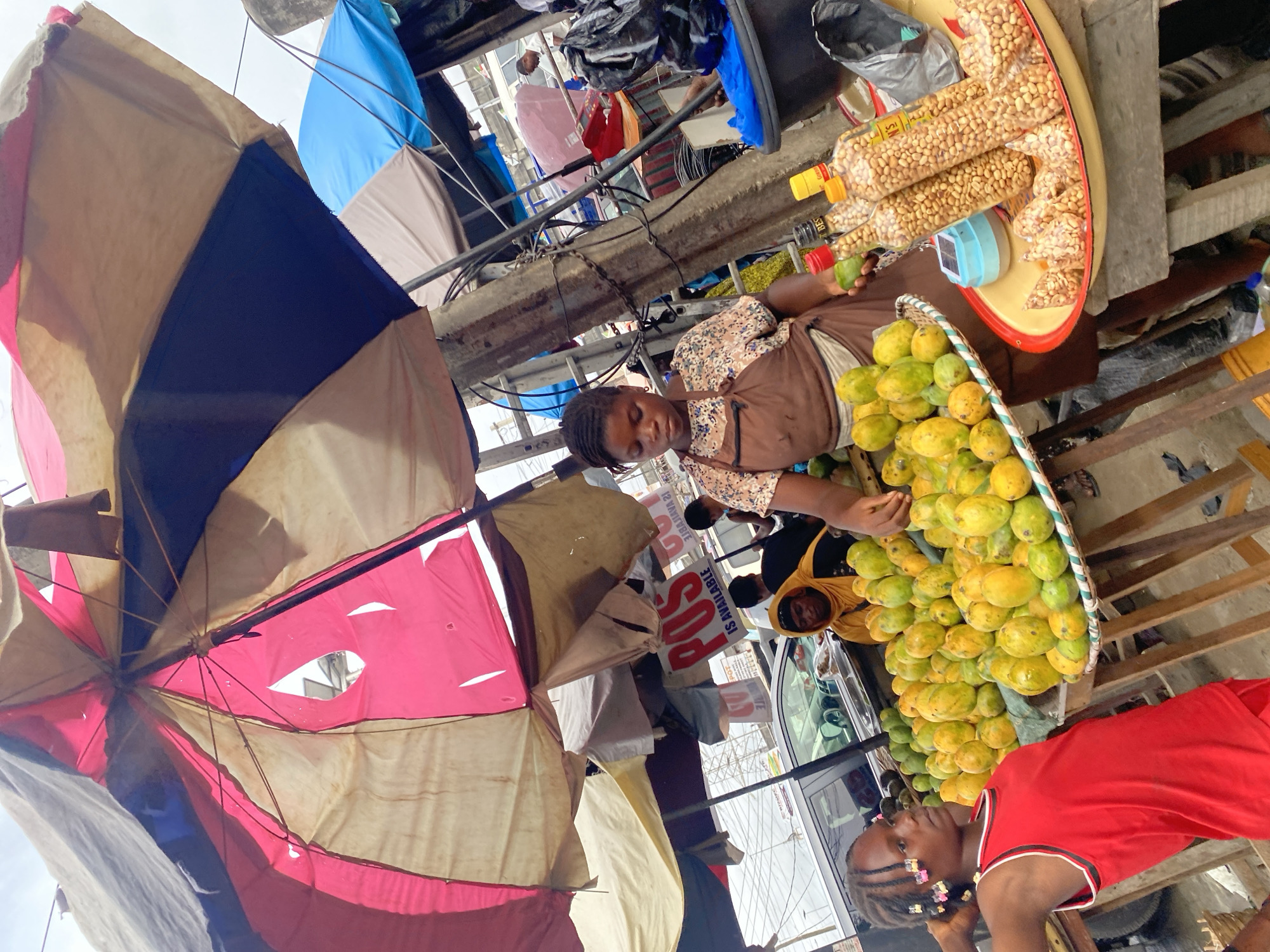 An eye-level, slightly rotated outdoor shot reveals a busy street scene in Lagos, Nigeria.
The scene is bustling with activity centered around a street vendor selling fruits and peanuts. A young woman in a patterned blouse and brown vest stands behind a large basket overflowing with green and yellow mangoes. To her right, a round yellow tray displays bottled peanuts. Above, an umbrella shades her stand. Another figure in a yellow hooded top is present near the fruit. A second child wears a red shirt.
The environment includes other vendors and their umbrellas. Behind the vendor, utility poles are visible, intertwined with wires, adding to the cluttered feel of the street. A white vehicle partially obscures the background. A sign with the letters "POS" is visible.