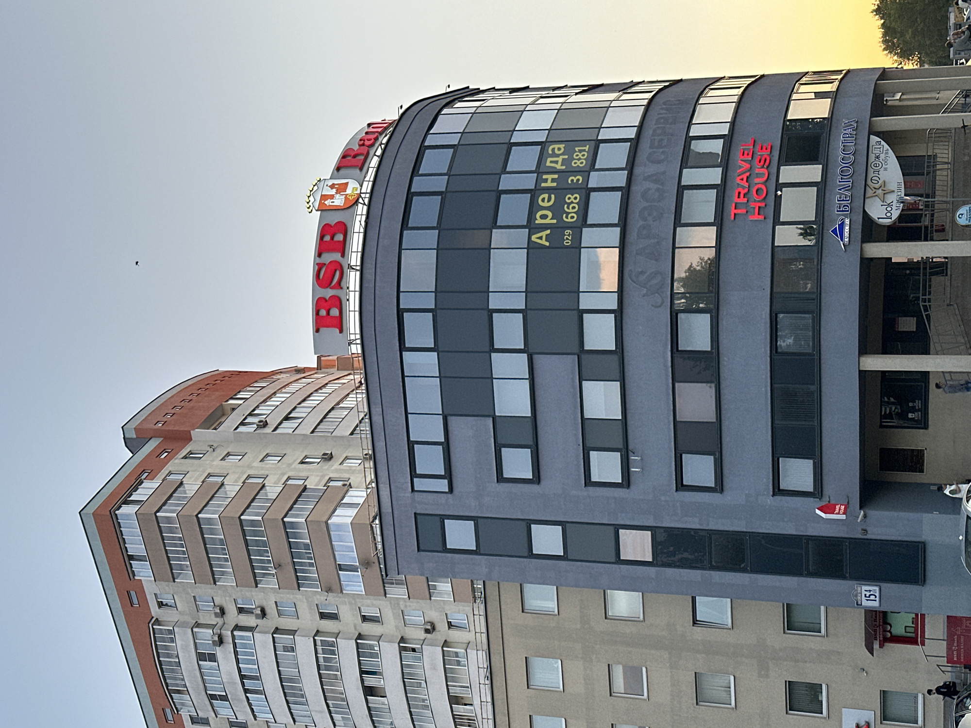 A late afternoon or early evening scene captures an urban streetscape featuring two prominent buildings under a clear sky. In the foreground, a modern, curved building presents a dark gray facade interspersed with numerous reflective windows. Prominently displayed at its top is a red "BSB Bank" sign, accompanied by a coat of arms logo. On the glass facade, yellow text reads "Аренда" (Rent) along with phone numbers "029 668 3 881". Faintly visible below is "АРЭСА СЕРВИС". Further to the right, red letters spell out "TRAVEL HOUSE". Adjacent to this, "БЕЛГОССТРАХ" (Belarusian State Insurance) is written, with smaller signs beneath for "look", "Одежда и обувь" (Clothes and shoes), and "магазин" (store). A small red arrow sign, vertically oriented with "TRAVEL HOUSE", is also present. A street sign displaying "154" is visible near the bottom.
Behind the modern structure stands a taller residential building, characterized by a wavy architectural design, numerous balconies, and a facade comprising tan, red-orange, and light gray panels. The sky above transitions from a pale blue to a warm, yellowish glow on the right, suggesting sunset or sunrise. A tiny, dark speck, possibly a bird, is seen high in the clear sky. Partial views of a vehicle and indistinct human figures are discernible at the street level near the base of the buildings. No specific activity or event is taking place; the image is a static portrayal of an urban setting.