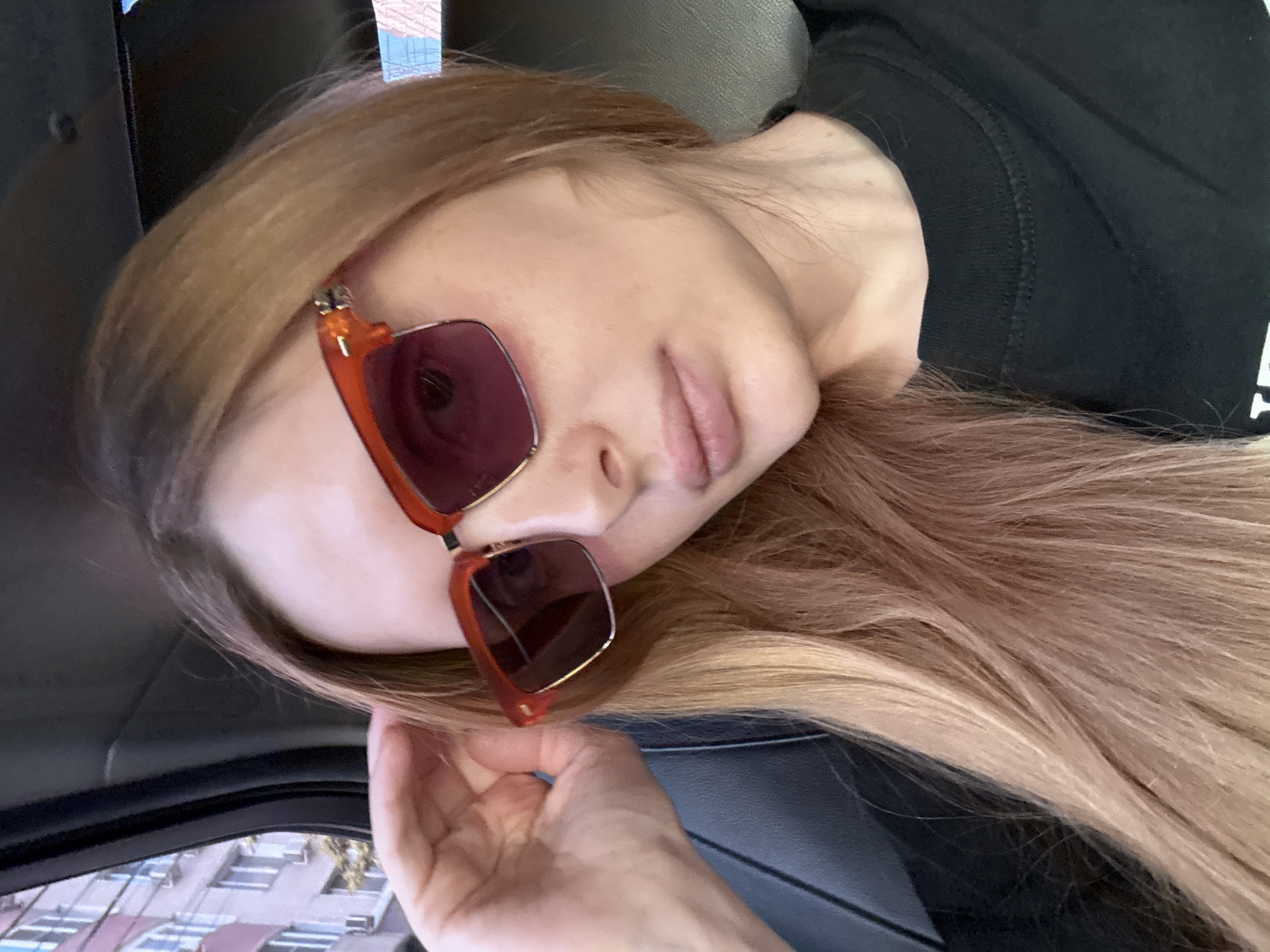 This media file features a close-up, horizontal shot of a young woman lying down, possibly in a car due to the visible car interior elements like the dark upholstery and window frame. She has light brown hair and is wearing a black top and distinctive orange-rimmed sunglasses with dark lenses. Her face is turned away from the camera, with her eyes obscured by the sunglasses. Her lips are slightly parted, and her hand is resting near her face, touching her hair. The background shows a blurred view of buildings through what appears to be a car window, suggesting an urban setting. The lighting suggests daytime, possibly with soft, diffused light, and the overall mood is relaxed or contemplative. No text is visible in the image. The location context provided is Krasnodar, Russia.