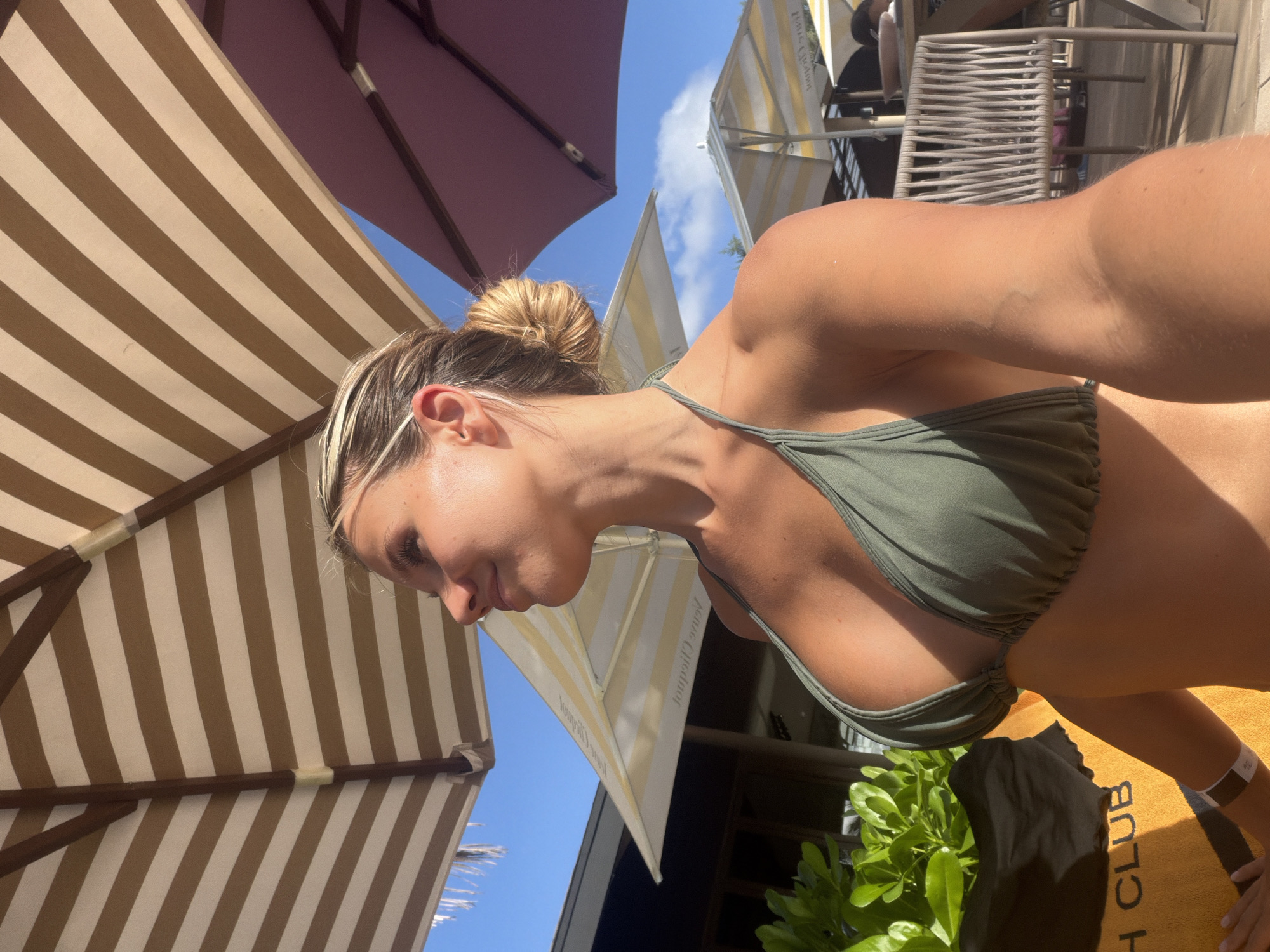 A young Caucasian woman is posing for a selfie outdoors under striped umbrellas, possibly at a resort in Choeng Thale, Thailand. She is wearing a green bikini top and has her blonde hair pulled up in a bun. Her head is turned to the side, and she is smiling. In the background, there are more umbrellas, lounge chairs, and lush green foliage. The sun is shining, casting strong shadows.