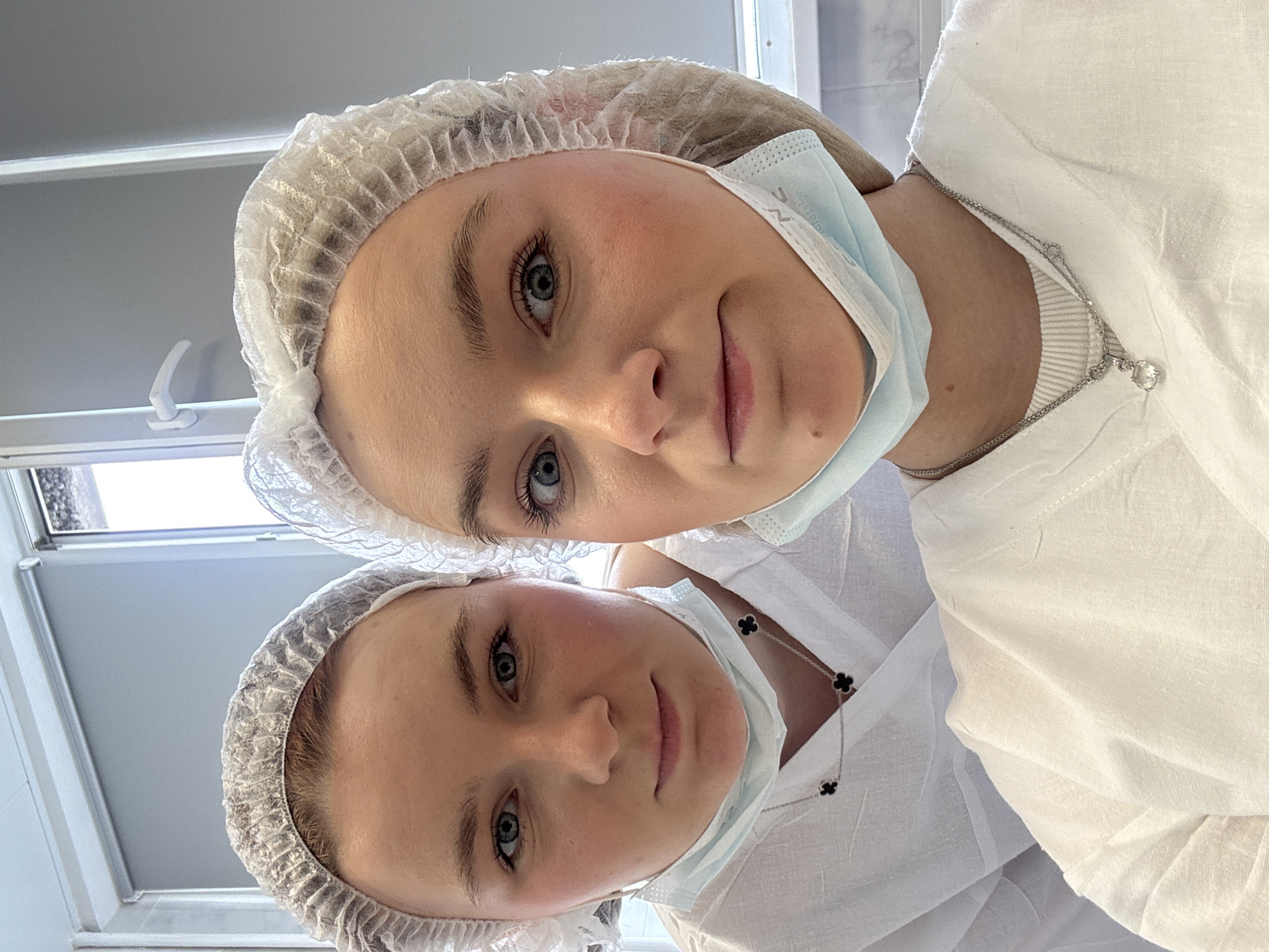 Two young women wearing white surgical caps and face masks, and white linen-like clothing, are looking down towards the camera. The woman in the top half of the frame has a silver necklace with a small pendant. The woman in the bottom half of the frame has a silver chain with black cloverleaf pendants. A window with a partially obscured cityscape visible outside is in the background on the top left of the frame. The location is possibly in an indoor setting in Saint Petersburg, Russia, based on the cityscape visible through the window.
