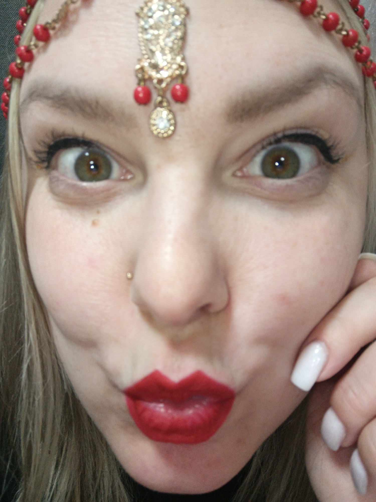 A close-up selfie shows a woman with fair skin, brown-green eyes, and blonde hair. She is wearing a decorative headpiece with red beads and a gold-colored pendant on her forehead. She has red lipstick and a nose piercing on her left nostril. Her left hand, with white manicured nails, is partially visible near her face. The location is not discernible from the image.