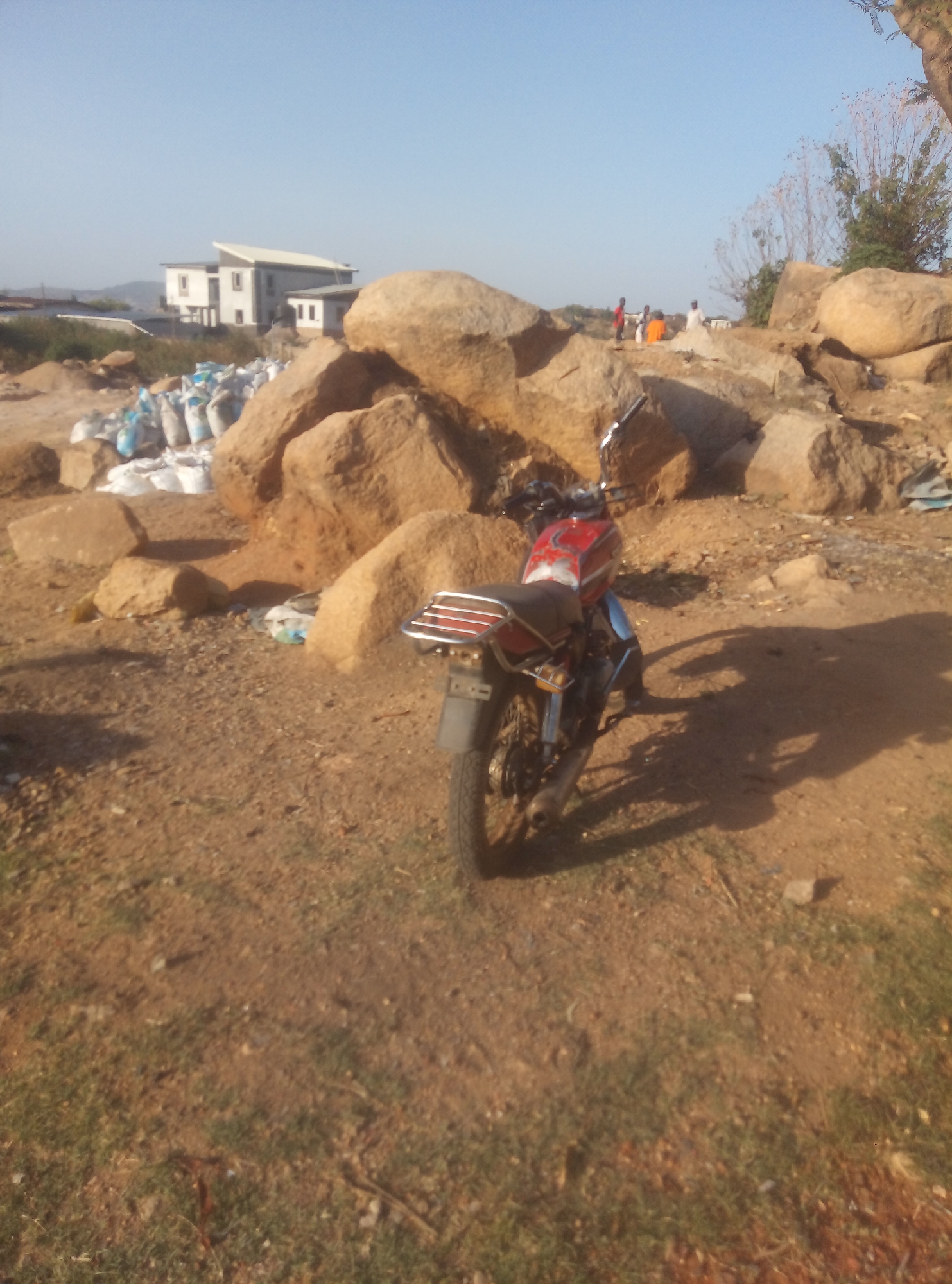 The image shows a red and blue motorcycle parked on a dirt and gravel surface, likely outdoors. Behind the motorcycle are several large, light-brown rocks and some white bags filled with an unidentified substance. In the background, there is a white, modern-style building and some figures standing near the rocks. The environment suggests a construction or industrial site located on a sunny day.