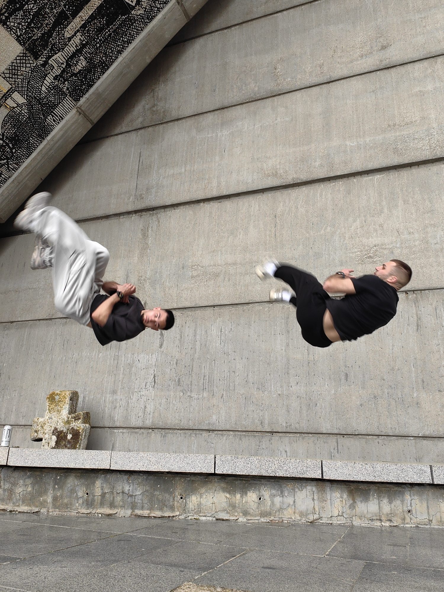 The image depicts two young men in mid-air, simultaneously performing acrobatic flips or parkour moves. The man on the left wears light gray pants and a dark t-shirt, with his arms crossed and knees bent. His face is visible, looking downwards. The man on the right is dressed in black pants and a black t-shirt, also with arms crossed and knees bent, and his face is visible, looking downwards.
They are set against a large, light grey concrete wall with distinct horizontal lines, suggesting pre-cast panels. An angled overhead structure features an intricate, dark mosaic-like pattern. The ground level includes a speckled granite ledge or bench, below which is a textured concrete base with small holes and cracks. The foreground shows grey paved ground.
On the ground near the left side, there is a weathered, moss-covered stone object shaped like a cross, and a white and silver can, partially visible, which appears to be a Monster Energy drink. The scene is brightly lit, indicating daytime. The environment suggests an urban or architectural space, possibly under an overpass or within a brutalist design structure, serving as a dynamic backdrop for the athletic activity.