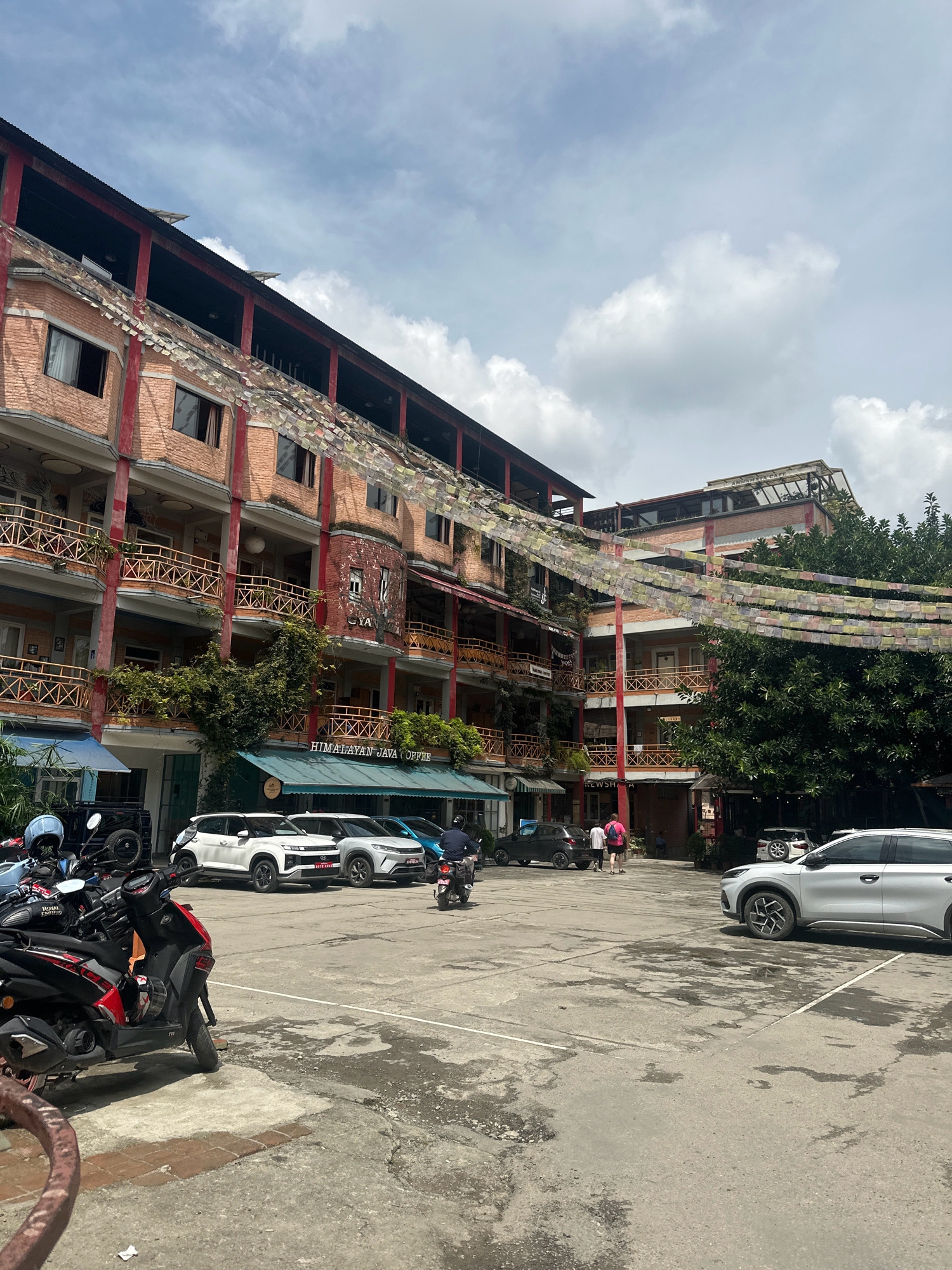 The image depicts the exterior of the Himalayan Java Coffee shop and Anamaya Cafe in Lalitpur, Nepal, with a view of a courtyard and several vehicles. The multi-story building features a brick facade with red columns and balconies adorned with traditional prayer flags strung across. Lush green foliage is visible, integrated into the building's architecture. In the foreground, several motorcycles are parked, with one rider on a motorcycle in the mid-ground. In the courtyard, several cars are parked, including a white SUV, a silver SUV, and a dark-colored hatchback. Two individuals are walking in the courtyard, and a few other people are seen in the background, near the buildings. The sky is partly cloudy.
