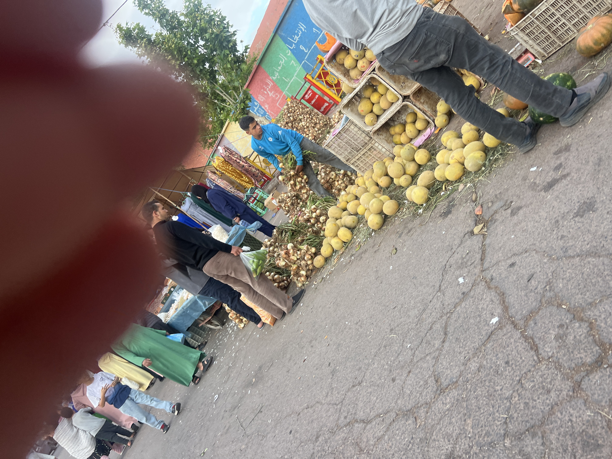 The image shows an outdoor market scene, likely in Ouaouizert, Morocco. A vendor in a blue shirt is handling onions next to a colorful wall with Arabic script. Several people, including a person in a grey shirt and pants, are standing near crates of yellow fruits and pumpkins. Other people are wearing traditional Moroccan clothing. The scene is on a paved surface with cracks. A hand is partially obstructing the view.