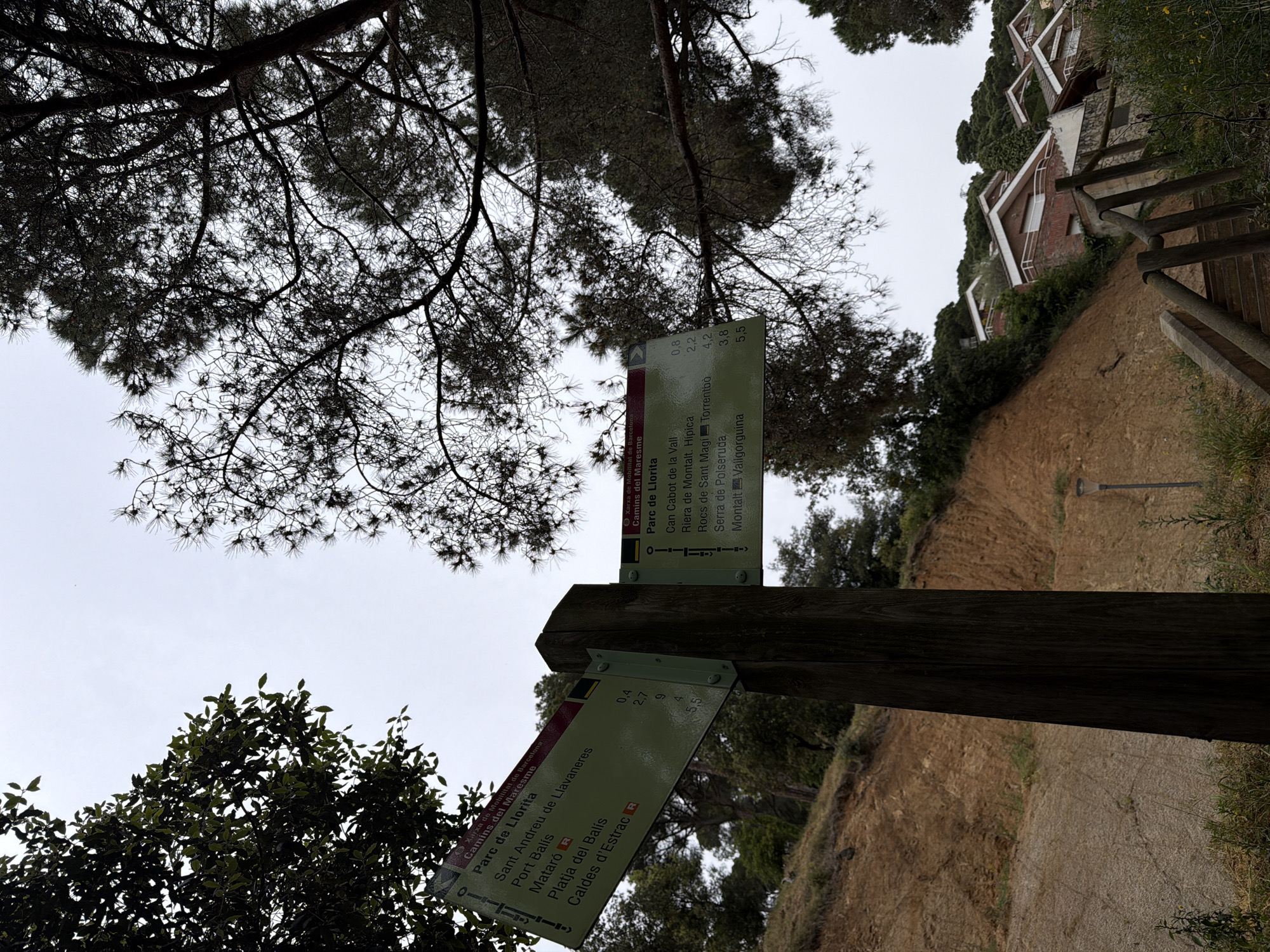 The image depicts a wooden signpost in Sant Andreu de Llavaneres, Spain, under the shade of trees. Two signs are attached to the post. The top sign lists directions to Can Cabot de la Vall (0.8 km), Riera de Montalt/Hípica (2.2 km), Rocs de Sant Magí Torrentbò (4.2 km), Serra de Polseruda (3.8 km), and Montalt Valigorguina (5.5 km). The bottom sign points to Sant Andreu de Llavaneres (0.4 km), Port Balís (2.7 km), Platja del Balís (9 km), and Caldes d'Estrac (5.5 km).