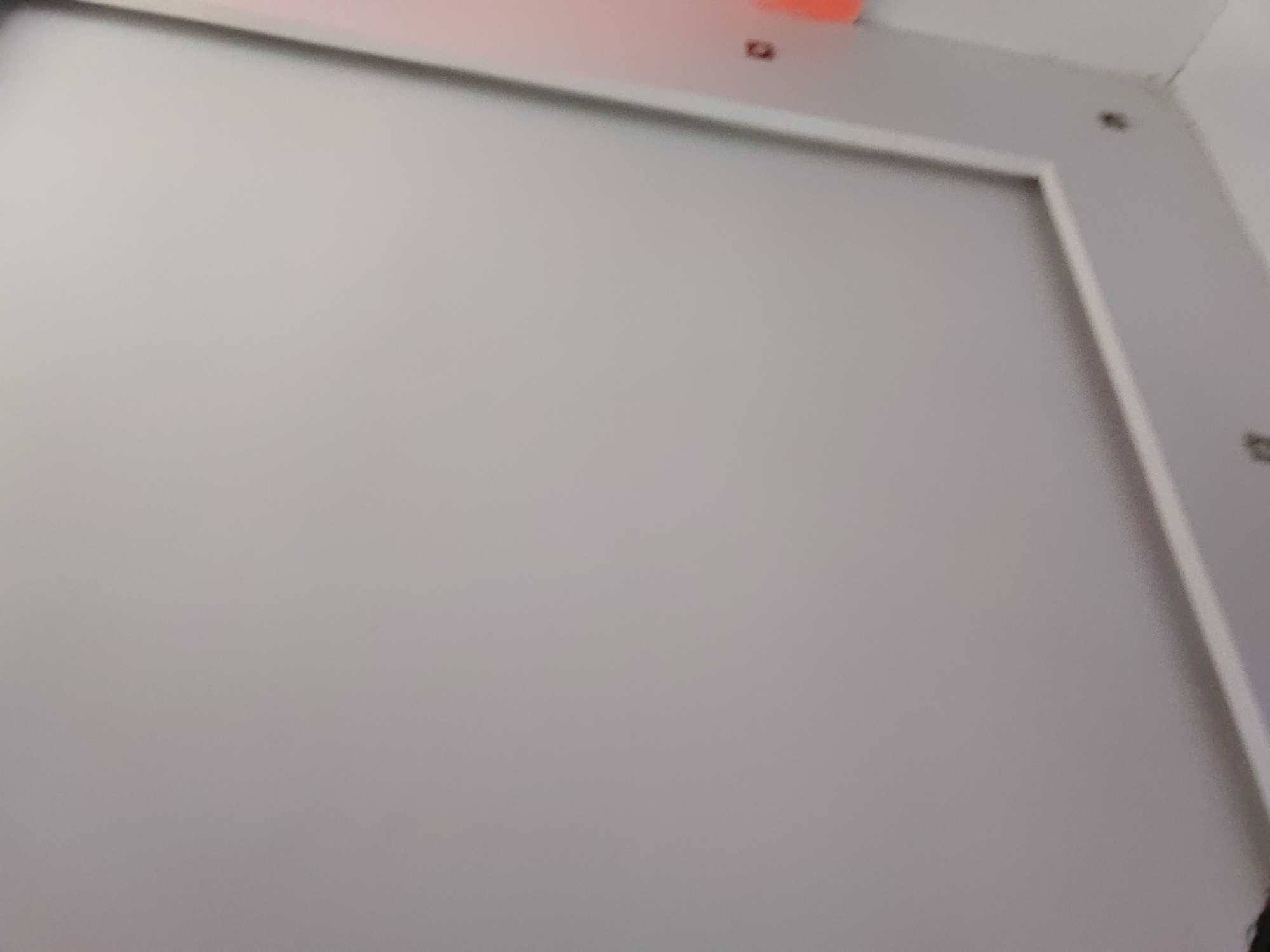 The image displays a close-up, angled shot of a light-colored, likely off-white or light gray, rectangular panel or door, which dominates the frame. This central surface is enclosed by a silver or metallic-colored frame, visible along the top, right, and partially the left edges.
In the upper central area, just above the metallic frame, a blurred orange or red object is partially visible, which could be an indicator light or a small fixture. Nearby, and also in the upper right corner of the frame, several small, dark, round objects are discernable, appearing to be screws or fasteners securing the frame or panel in place. The perspective suggests the photograph was taken from a slight upward or angled position.
No individuals are present, and no particular activity is depicted. The scene is static, showing an interior architectural or structural element. There are no direct visual cues within the image to specify the time of day, weather conditions, or to confirm the provided location context of Santa Ana, Venezuela. No text is visible in any part of the image. The overall impression is of a functional, minimalist surface within an indoor setting.