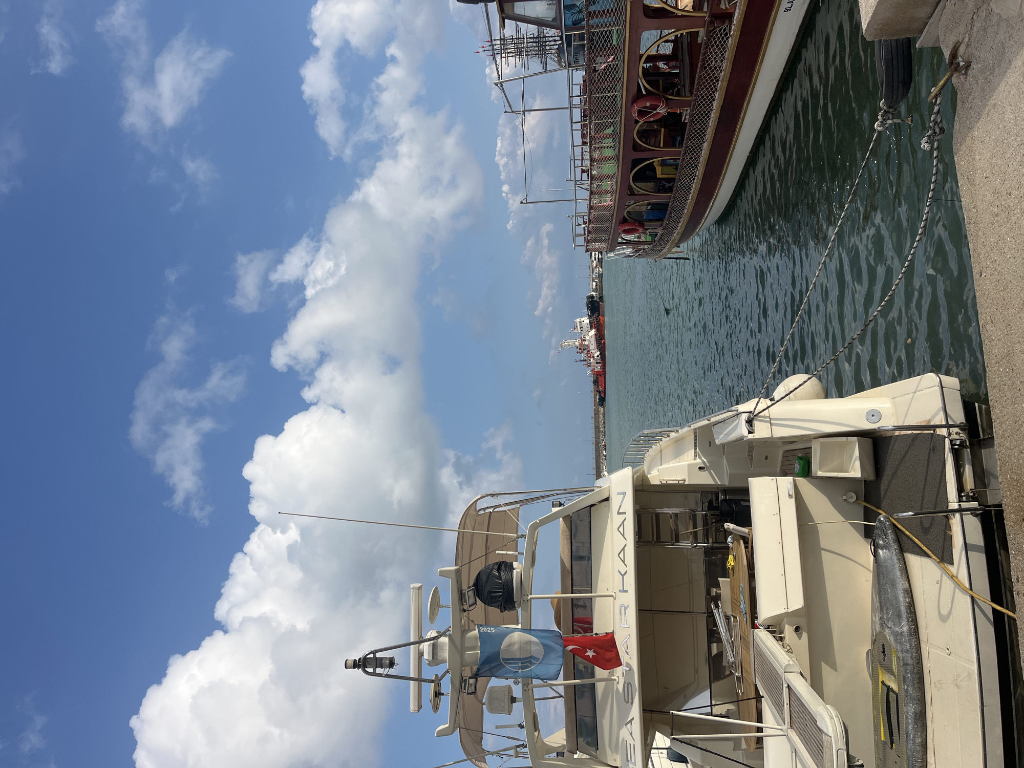 The image shows a marina in Alanya, Turkey, on a sunny day with fluffy white clouds against a clear blue sky. Two boats are prominently featured: a brown and white wooden boat docked alongside a modern, white yacht bearing the name "ARKASAAR KAAN" on its stern. The yacht also has a flag with a symbol and the year 2025 along with a Turkish flag. In the background, several additional vessels are visible at the harbor. The water in the marina appears calm, reflecting the sky.