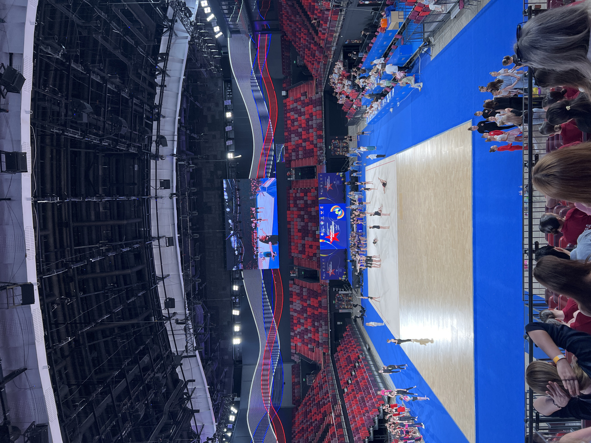 This is a wide shot of a gymnastics competition taking place in a large indoor arena. The arena has tiered seating for spectators, with rows of red and purple seats visible. The competition floor is a light-colored mat surrounded by a blue border. Several young gymnasts are on the floor, some performing routines and others in groups. There are also officials and coaches present. Above the competition area, a large video screen displays footage of a gymnastics event. The arena ceiling is complex, with numerous lights, speakers, and structural elements suspended from it. Red, white, and blue decorative lights are strung along the upper part of the arena walls. The scene appears to be at the Luzhniki Palace of Gymnastics in Moscow, Russia, as evidenced by the signage visible on the large screen and banners.