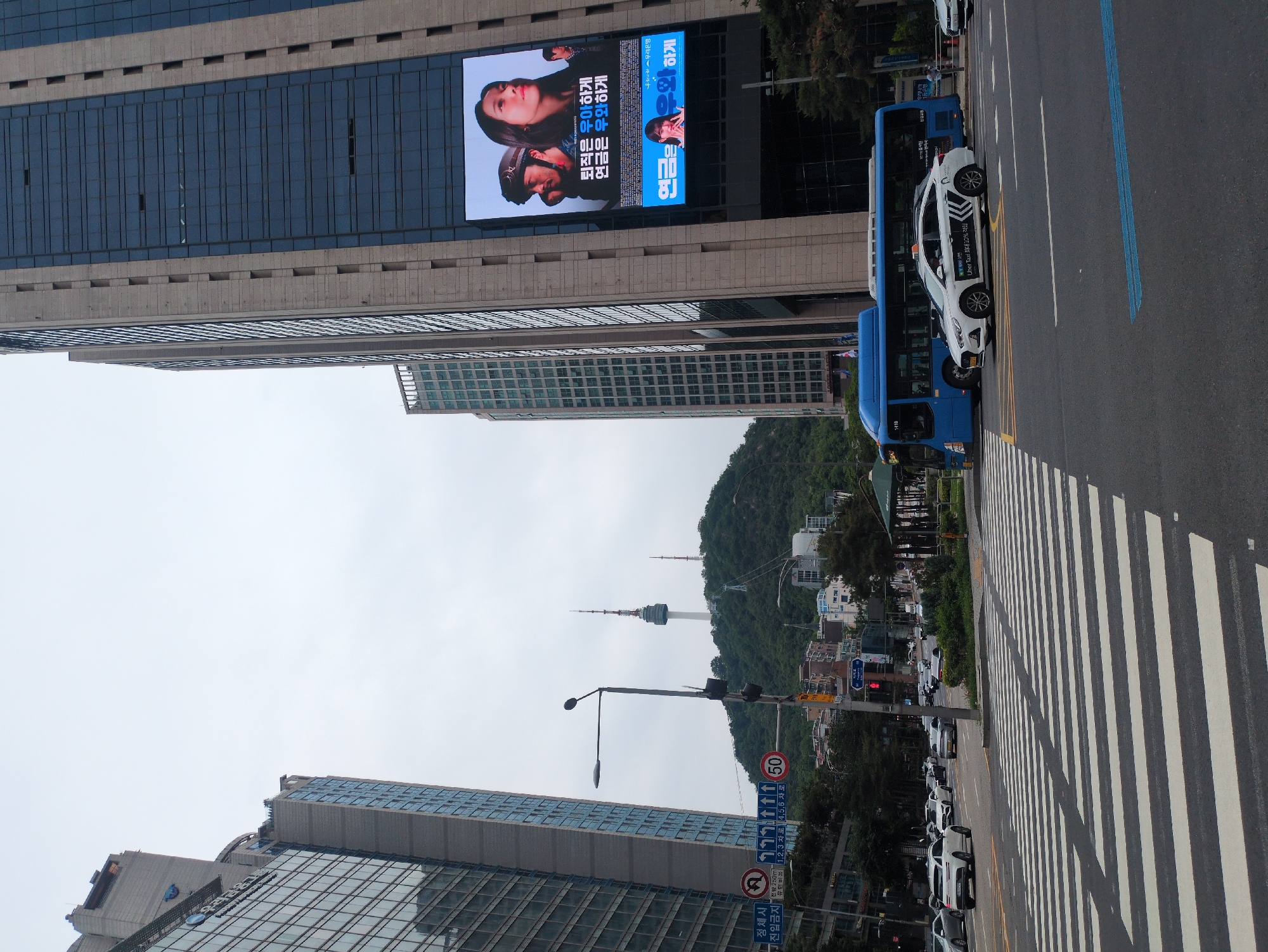 This image captures a busy urban street scene in Seoul, South Korea, facing south-southeast on Namdaemun-ro in Jung-gu, with Namsan Tower visible on a forested hill in the background. The sky is uniformly overcast.
In the foreground, a wide asphalt road features a prominent diagonal pedestrian crosswalk with alternating thick white and black stripes. A white taxi, identified as an "Uber Taxi" by text on its side, is positioned near the crosswalk. Behind it, a blue city bus with "1410" displayed on its front approaches or is stopped. Further down the road, several cars, including white, gray, and silver sedans and SUVs, are visible in multiple lanes, some appearing to be waiting at a distant intersection with a red traffic light. A blue line is marked on the asphalt in the lower right. A black street light pole with multiple traffic signals and CCTV cameras stands on the left side of the street.
In the midground, traffic signs are visible, including a red circular speed limit sign indicating "50" km/h, a blue and white sign with Korean text "정체시 전방 250m 유턴위반" (Congestion: U-turn violation 250m ahead) and "1,2,3,4,5,6 차로" (Lanes 1,2,3,4,5,6), and a "No Right Turn" sign. A green-canopied shelter, possibly a bus stop, is situated on the right, surrounded by small trees and shrubs.
The background is dominated by a densely forested hill, Namsan, upon which Namsan Tower (N Seoul Tower) stands prominently. The tower is white with a distinctive antenna spire. A smaller communication tower is also visible to its right. Buildings are scattered across the lower slopes of Namsan.
Framing the scene are tall, modern high-rise buildings. The building on the left features a glass facade with reflections and gray stone cladding; a blue logo and Korean text identifying it as "BNK 부산은행" (Busan Bank) are visible near its top. The building on the right is similarly tall, constructed with dark glass windows and light-colored stone cladding. A large, bright digital billboard is integrated into its facade. This billboard displays an advertisement for "우리은행" (Woori Bank), featuring images of a woman looking upwards and a man in a helmet, with Korean text reading "퇴직은 우아하게" (Retirement elegantly) and "연금은 우아하게" (Pension elegantly). A smaller section below repeats the latter phrase with another female figure.