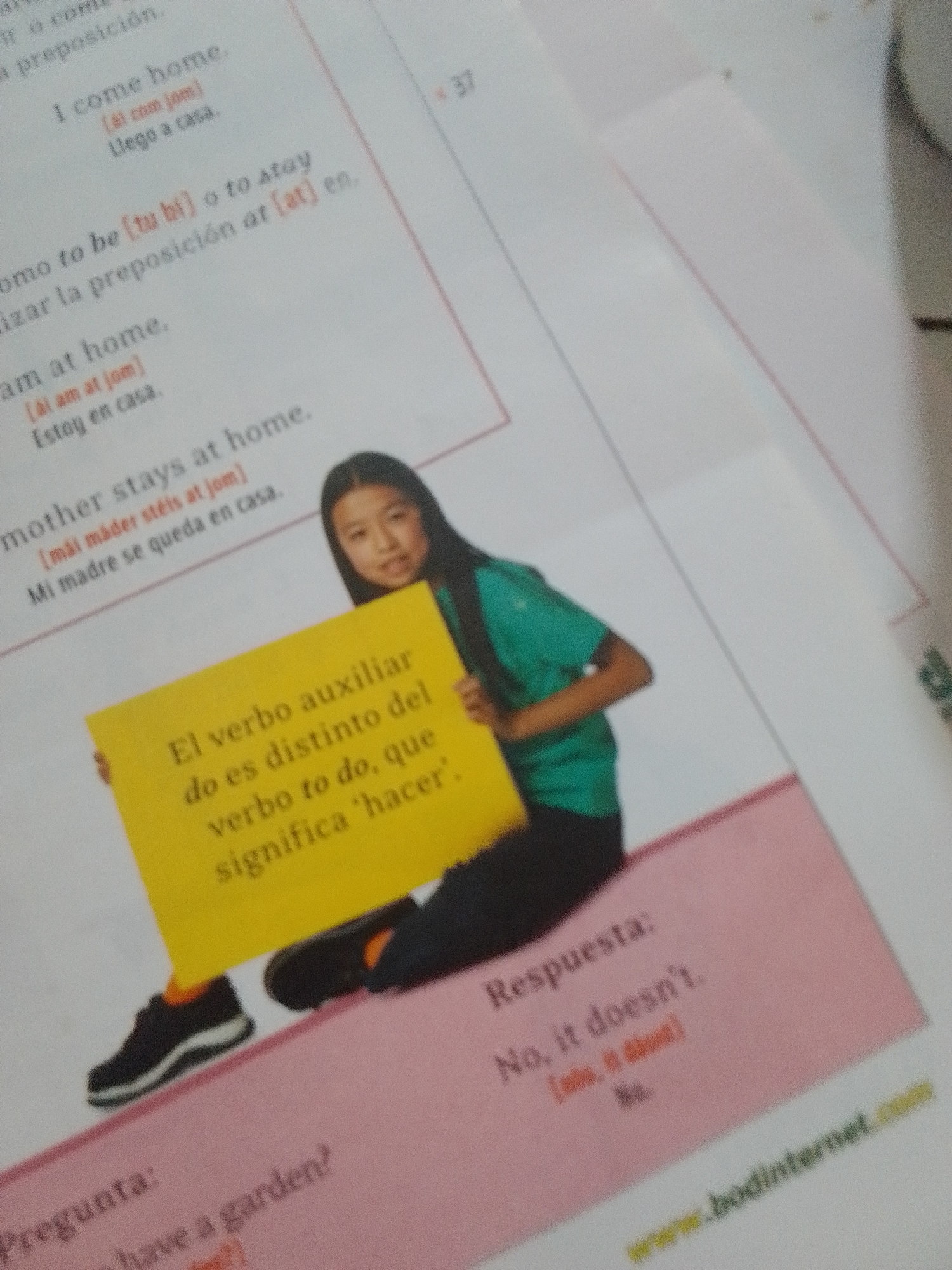 This media file captures a close-up of a book or workbook, likely in a classroom or study setting, with a young person holding a yellow sign. The scene appears to be indoors, as indicated by the lighting and the presence of printed educational material.
The primary subject is a young person, likely a student, who is partially visible from the shoulders down, wearing a green shirt and dark pants. They are holding up a bright yellow sign with text written in Portuguese. The sign's content appears to be related to language learning, specifically concerning verbs and their meanings. Visible text on the sign reads "El verbo auxiliar do es distinto del verbo to do, que significa 'hacer'."
The book itself displays text in Portuguese and English, suggesting a bilingual educational context. Several lines of text are visible, including phrases like "I come home," "am at home," and "mother stays at home," along with their Portuguese translations and phonetic spellings. There are also sections labeled "Pregunta:" (Question:) and "Respuesta:" (Answer:), indicating an exercise or lesson format. The answer provided is "No, it doesn't." A website address, "www.bodinternet.com," is also visible at the bottom right of the frame.
The overall impression is one of learning and instruction. The image is well-lit, though the angle suggests it was taken with a mobile device from above. There are no strong cues about the time of day or weather, and no overt emotional expressions are visible due to the framing of the subject. The location context of La Tagua, Venezuela, is not explicitly indicated within the image itself, but if provided, it suggests the setting for this educational activity.