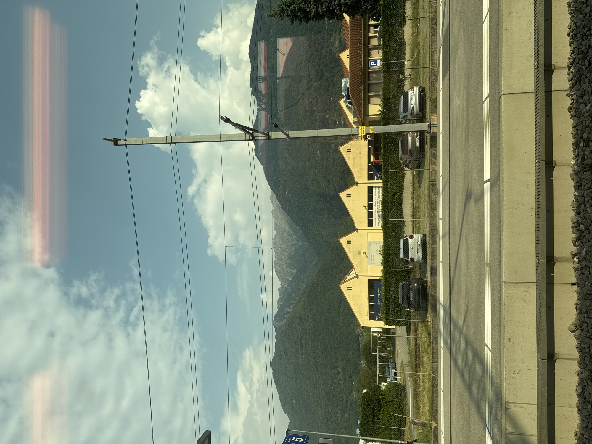 The image depicts a scenic view from a train window, likely in the vicinity of Bellinzona, Switzerland. The foreground features a train track with a concrete barrier and gravel. In the middle ground, a row of yellow buildings with distinctive triangular roofs, resembling warehouses or industrial units, is visible. Several parked cars, including a dark gray Volkswagen Golf, a silver Audi A3, and a white Volkswagen Golf, are parked in front of these buildings. Beyond the buildings, a dense, green, tree-covered mountain range dominates the background, with its peak partially obscured by clouds. A tall, gray utility pole with electrical wires runs vertically through the center of the frame. The sky is a clear blue with scattered white clouds. A blue sign with the number "5" is mounted on a pole on the left side of the image, possibly indicating a platform or bus stop. The overall scene is one of a functional industrial area set against a dramatic natural landscape.