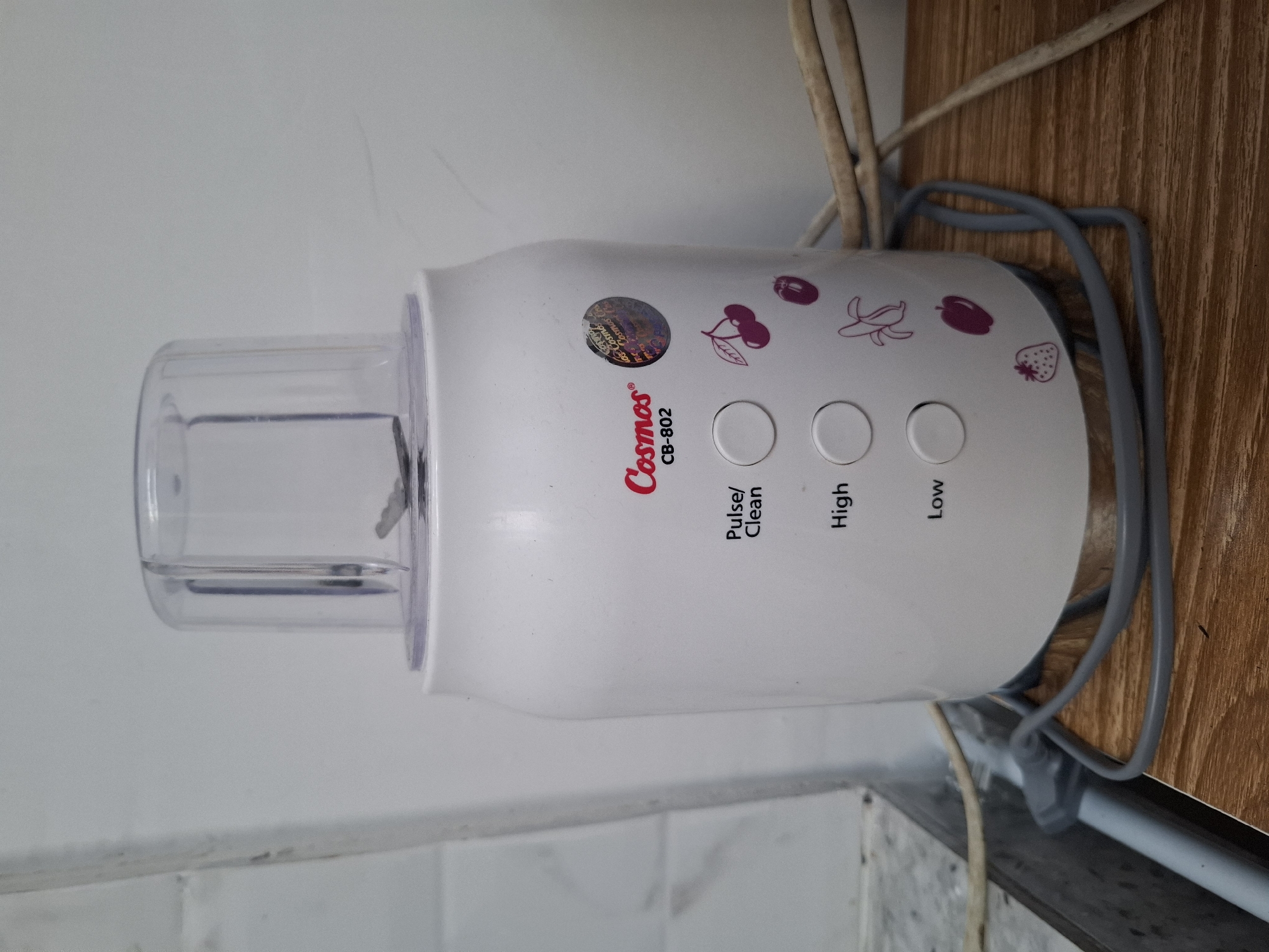 A white Cosmos CB-802 blender with a clear lid sits on a wooden surface against a white wall. The blender has three circular buttons labeled "Pulse/Clean," "High," and "Low." Decorative fruit illustrations, including cherries, a plum, a banana, an apple, and a strawberry, are printed on the blender's body. A power cord is plugged into the blender and lies on the wooden surface. The scene is likely in a kitchen or food preparation area within a home in Hulaan, Indonesia.