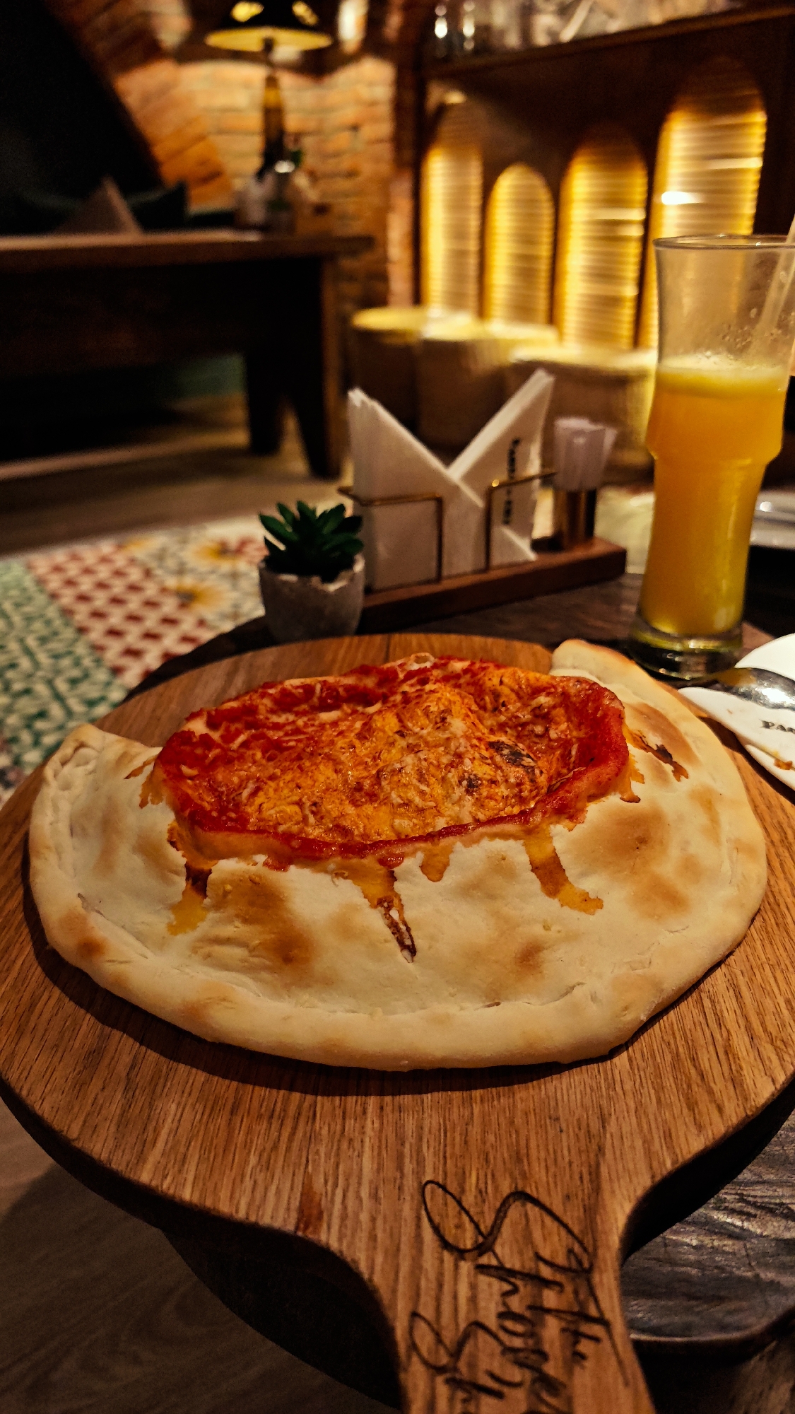A close-up shot shows a calzone pizza topped with tomato sauce and cheese, served on a wooden board. To the right, a tall glass of orange juice with a straw is visible. In the background, a rustic brick wall and warm lighting create an inviting atmosphere. The scene appears to be set in a restaurant, possibly in Hanoi, Vietnam, given the hints of traditional Vietnamese decor in the patterned carpet and the restaurant's ambiance. The calzone's crust is golden brown and slightly puffed, with some cheese and sauce spilling over the edges. A small potted succulent plant adds a touch of greenery to the composition. The overall impression is one of comfort and delicious food in a cozy setting.