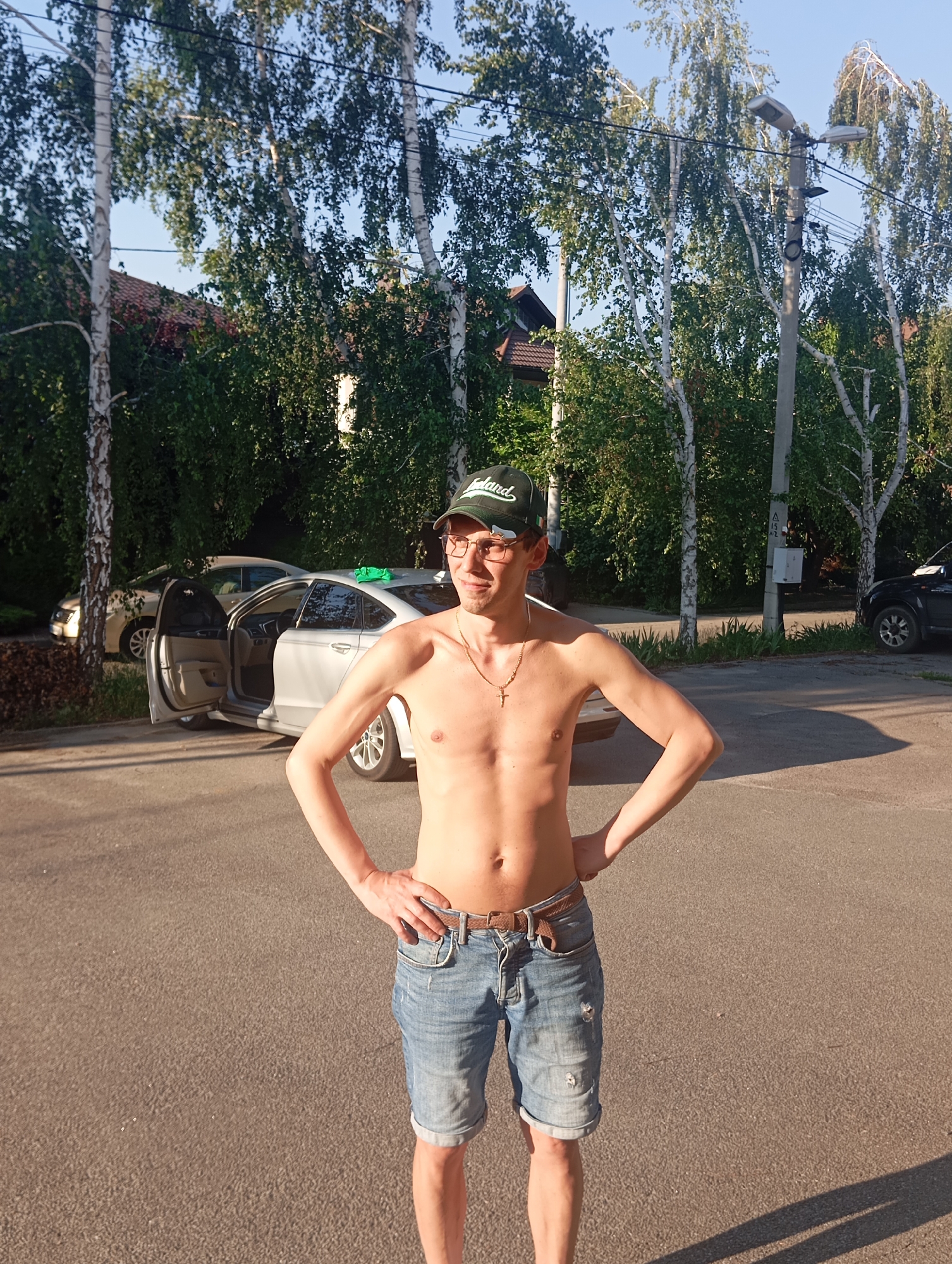 A shirtless young man with a lean build stands in a street in Sofiivska Borshchahivka, Ukraine. He is wearing denim shorts, a green baseball cap, glasses, and a gold necklace with a cross pendant. His hands are on his hips. In the background, a silver sedan with its driver's side door open is parked on the asphalt road. Another car is visible further down the street, and a black SUV is parked on the right side. The scene is bordered by trees and a building with a red tiled roof. A lamppost with two lights stands near the right edge of the frame. The ground is paved with asphalt, and sunlight casts shadows on the road.