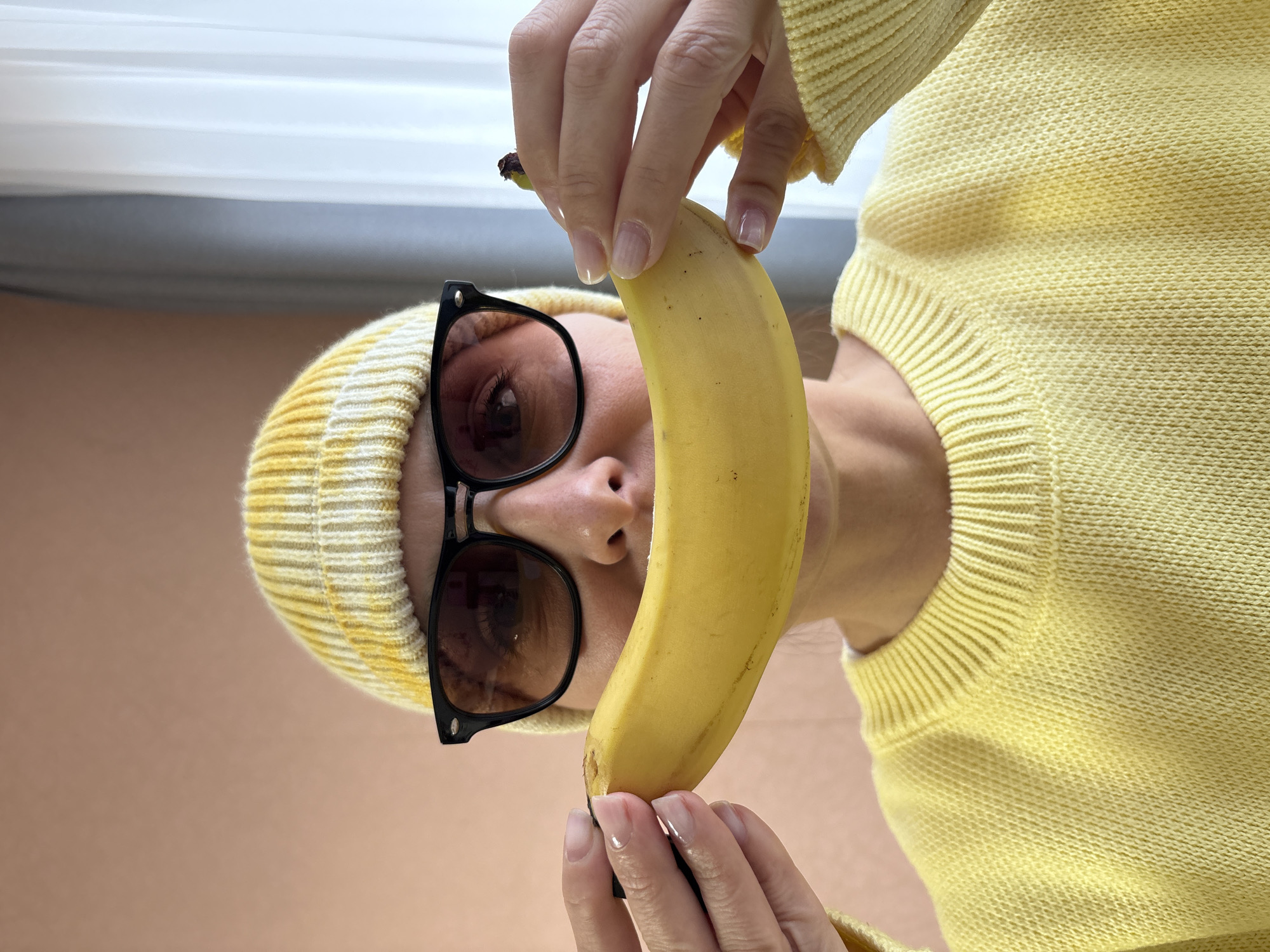 A person wearing a yellow sweater, yellow beanie, and dark sunglasses is holding a banana vertically in front of their face. The person's hands are holding the top and bottom of the banana. The background consists of an off-white window frame at the top and a beige wall at the bottom. The location, Chaikovsky, Russia, cannot be determined from the visual information.