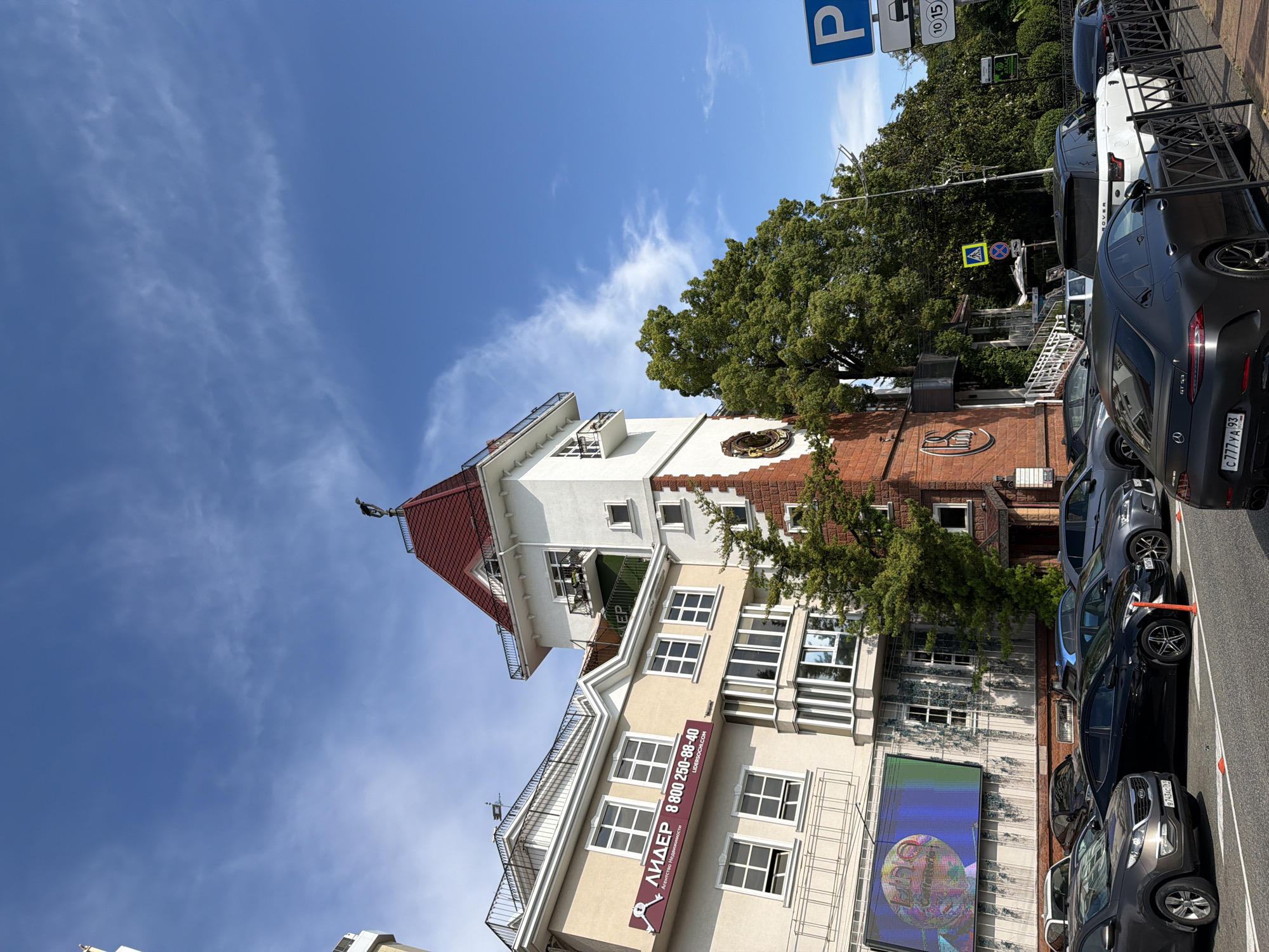 This is a medium-angle, rotated shot of a city street scene on a sunny day in Sochi, Russia. The primary subject is a unique, multi-story building with a distinctive red-tiled mansard roof and a white tower, accented by red brickwork. The building appears to be an office or commercial property, as evidenced by the signage. One prominent sign, in Russian, reads "ЛИДЕР" (LIDER) and displays a phone number "8 800 250-88-40", along with "Агентство Недвижимости" (Real Estate Agency) and a website "LIDERSOCHI.COM". Another sign features a stylized logo.
A row of parked cars lines the street in front of the building, including a black Mercedes-Benz with a license plate that reads "C777YA 93", and a white Range Rover. Overhead, a bright blue sky with wispy white clouds suggests fair weather. A blue parking sign is visible, indicating parking regulations. Tall, lush green trees partially obscure some of the building and provide a natural element to the urban setting. The overall impression is of a vibrant, bustling city environment. The image seems to capture an ordinary day, with no specific event taking place, but rather a snapshot of daily life in this Russian city.