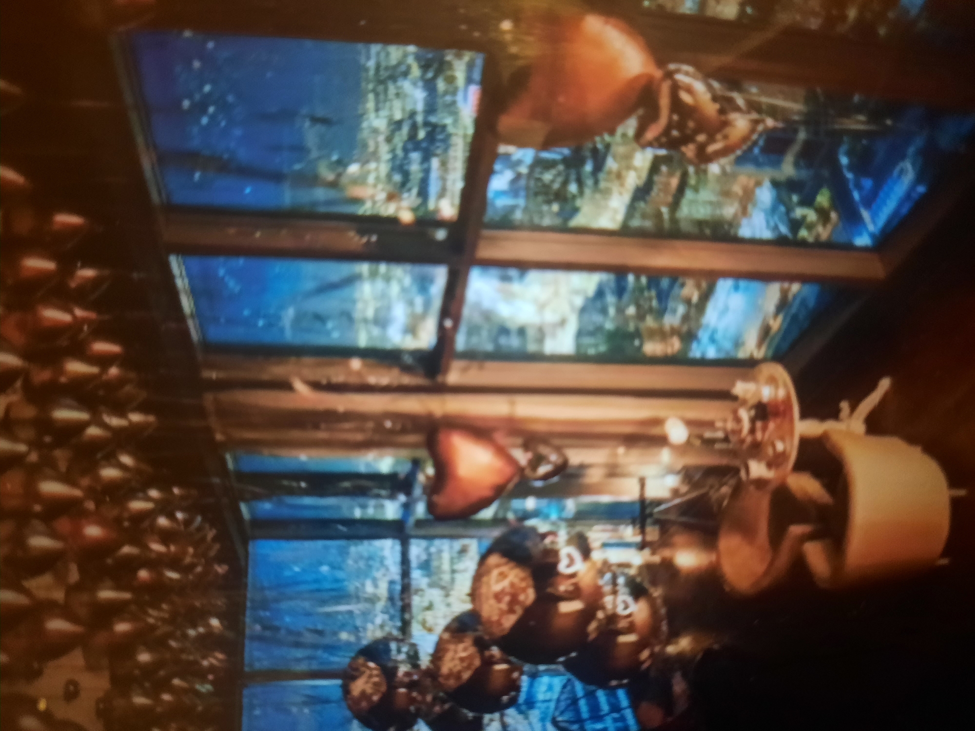 The image depicts an indoor scene with a view of a city at night through large windows. The room is decorated with numerous balloons, many of which are heart-shaped and metallic, with some clustered near the ceiling and others floating at various heights. In the foreground, there is a cream-colored armchair and a small round table with what appears to be some decorative items or a small centerpiece. The windows offer a panoramic view of a city illuminated by countless lights, suggesting a high vantage point. The lighting within the room is dim, creating a mood that is likely intended to be romantic or celebratory. The overall impression is one of a well-decorated private space overlooking a cityscape at dusk or night. The image does not provide any specific details to locate it in Rosenheim, Germany.