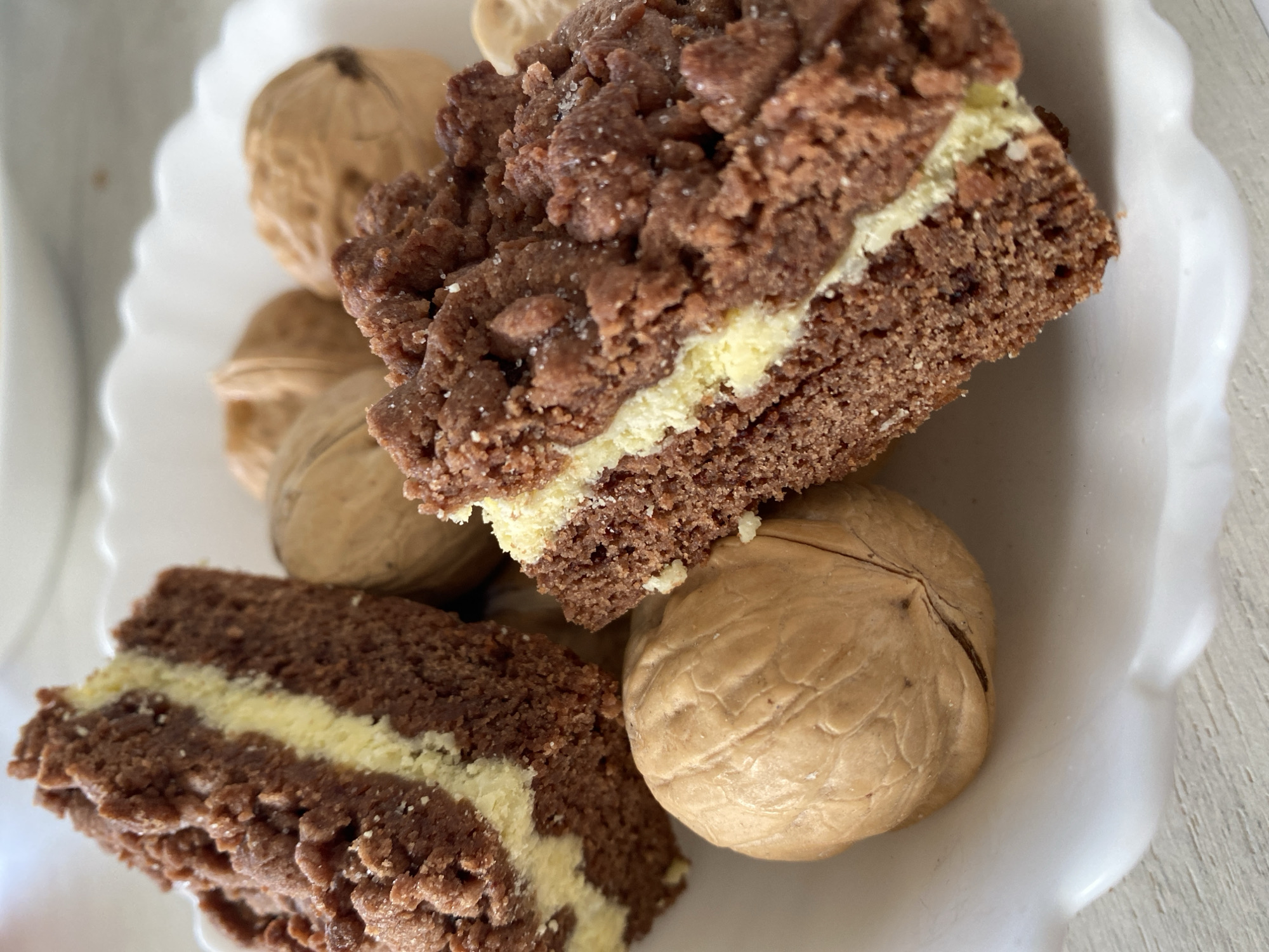 A close-up, overhead shot shows two pieces of chocolate cake with a cream filling, arranged on a white, scalloped-edge plate. Several whole walnuts are scattered around the cake pieces on the plate. The cake has a crumbly, textured topping and a dense, moist interior. The lighting is bright, suggesting an indoor setting. The background is out of focus, but appears to be a neutral-colored surface, possibly a table. There is no indication of location in Міханавіцкі сельскі Савет, Belarus.