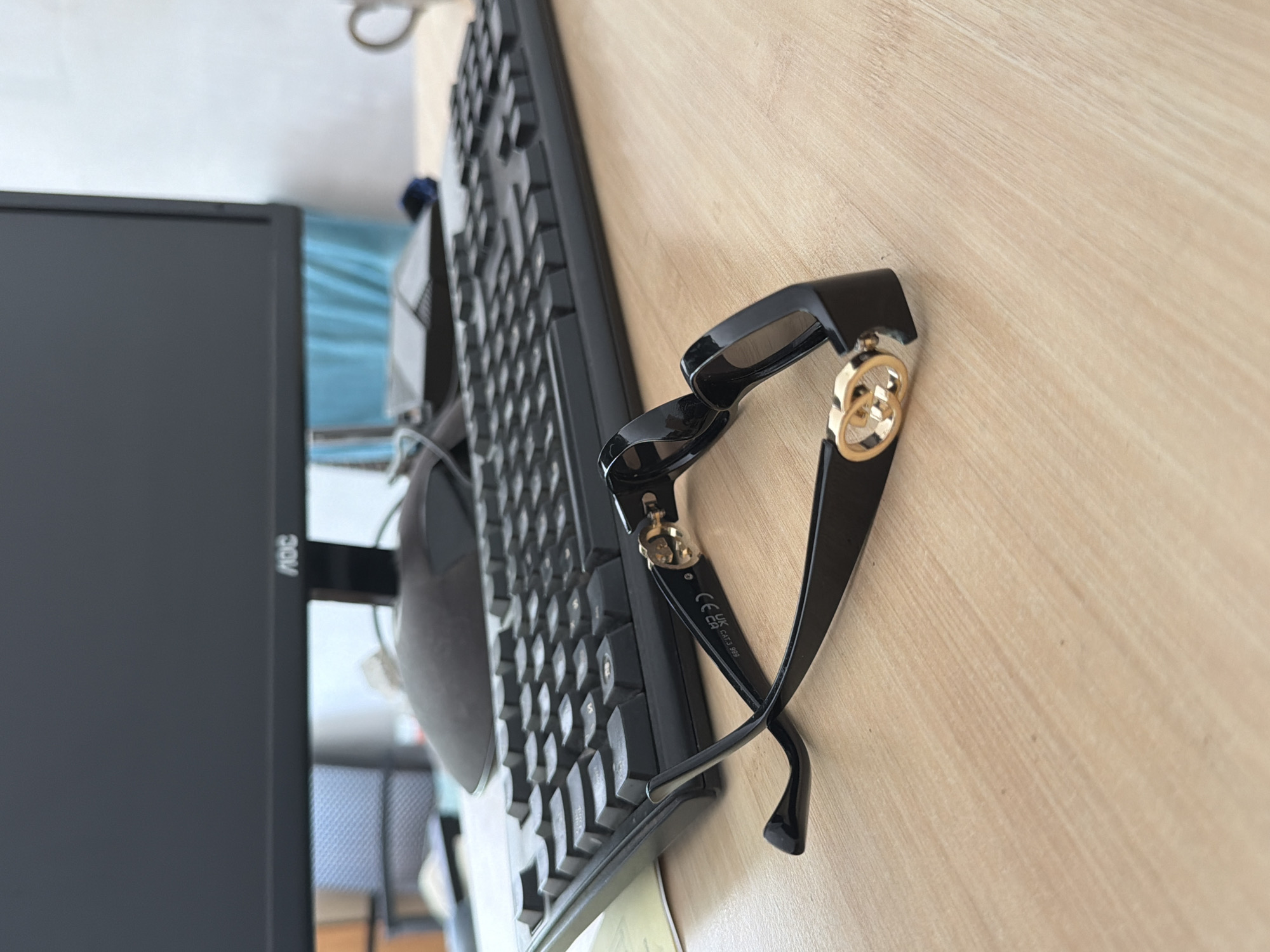 A pair of black-rimmed sunglasses with gold interlocking 'G' emblems rests on a light-colored wooden desk. The sunglasses are positioned diagonally, with the temples crossed. In the background, a black computer keyboard is visible, followed by a black computer monitor with the 'AOC' logo. To the right of the monitor, a white ceramic mug is partially visible. The overall environment suggests an office or workspace. The image does not contain any information that would suggest a location in Prosena, Bulgaria.