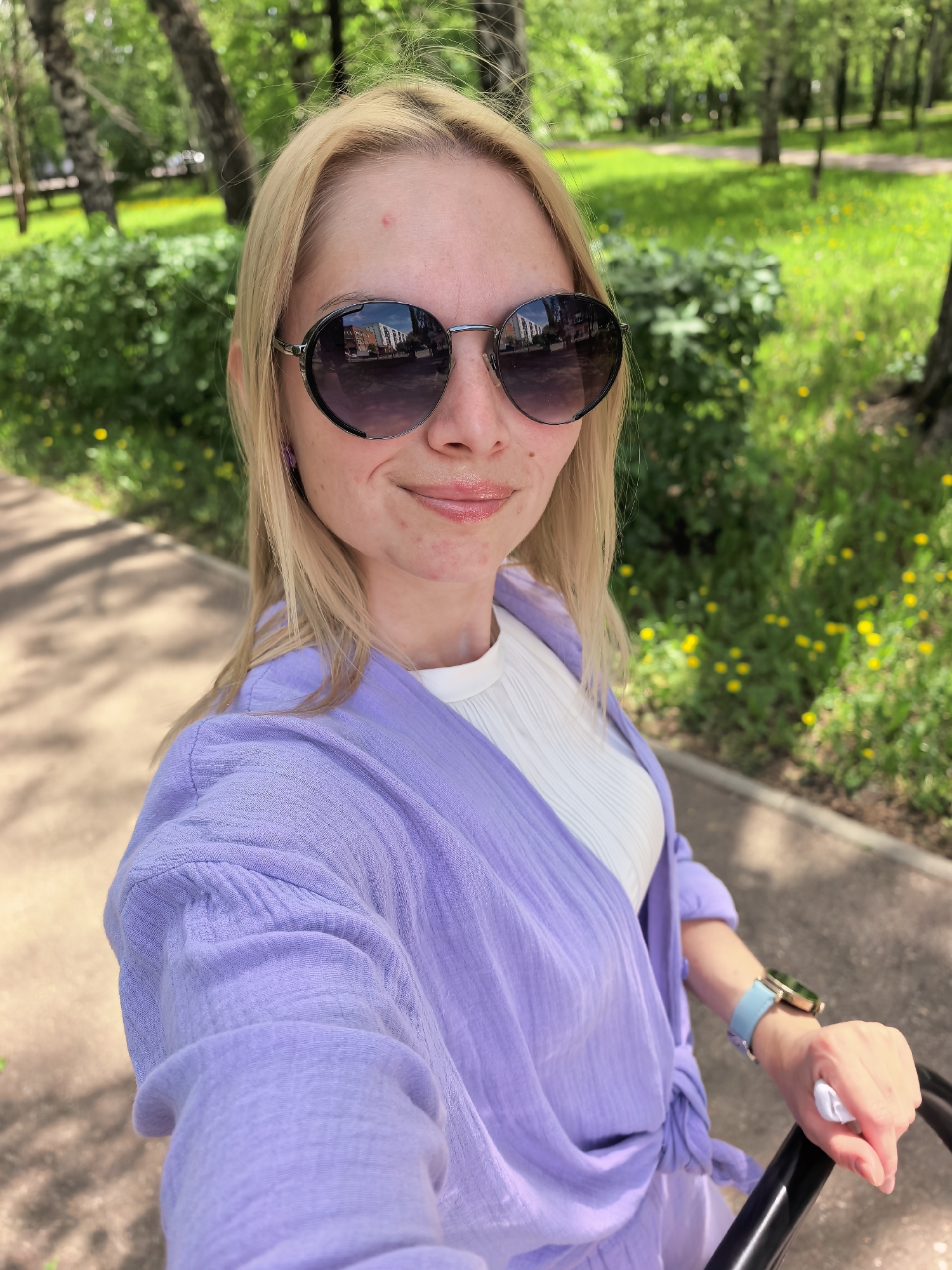 A fair-skinned woman with blonde hair, wearing sunglasses, a white shirt, and a lavender cardigan, is taking a selfie outdoors. She has a light blue watch on her left wrist. The sunglasses reflect buildings, suggesting an urban environment. The background shows a park with green grass, trees, and a pathway, indicating a city park setting. The photo was taken in Sterlitamak, Russia, as inferred from the reflection in the glasses of a building in the city. The woman is holding onto a black object, potentially part of a stroller or chair.