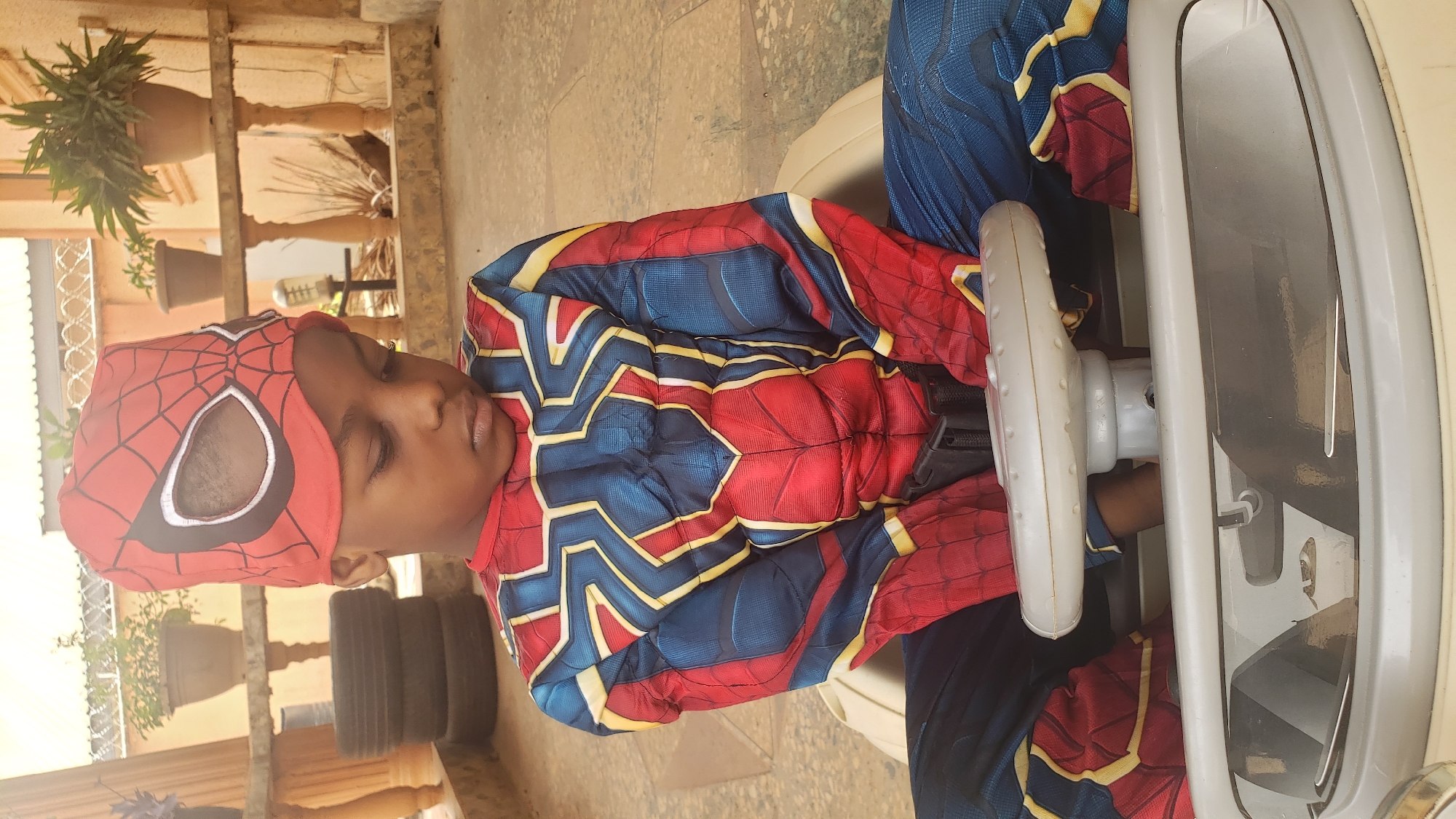 A child wearing a Spider-Man costume is seated in a white toy car with a steering wheel. The location appears to be outdoors, possibly on a patio or veranda, with potted plants and stacks of tires visible in the background. The image was captured in Sabon Tasha, Nigeria.