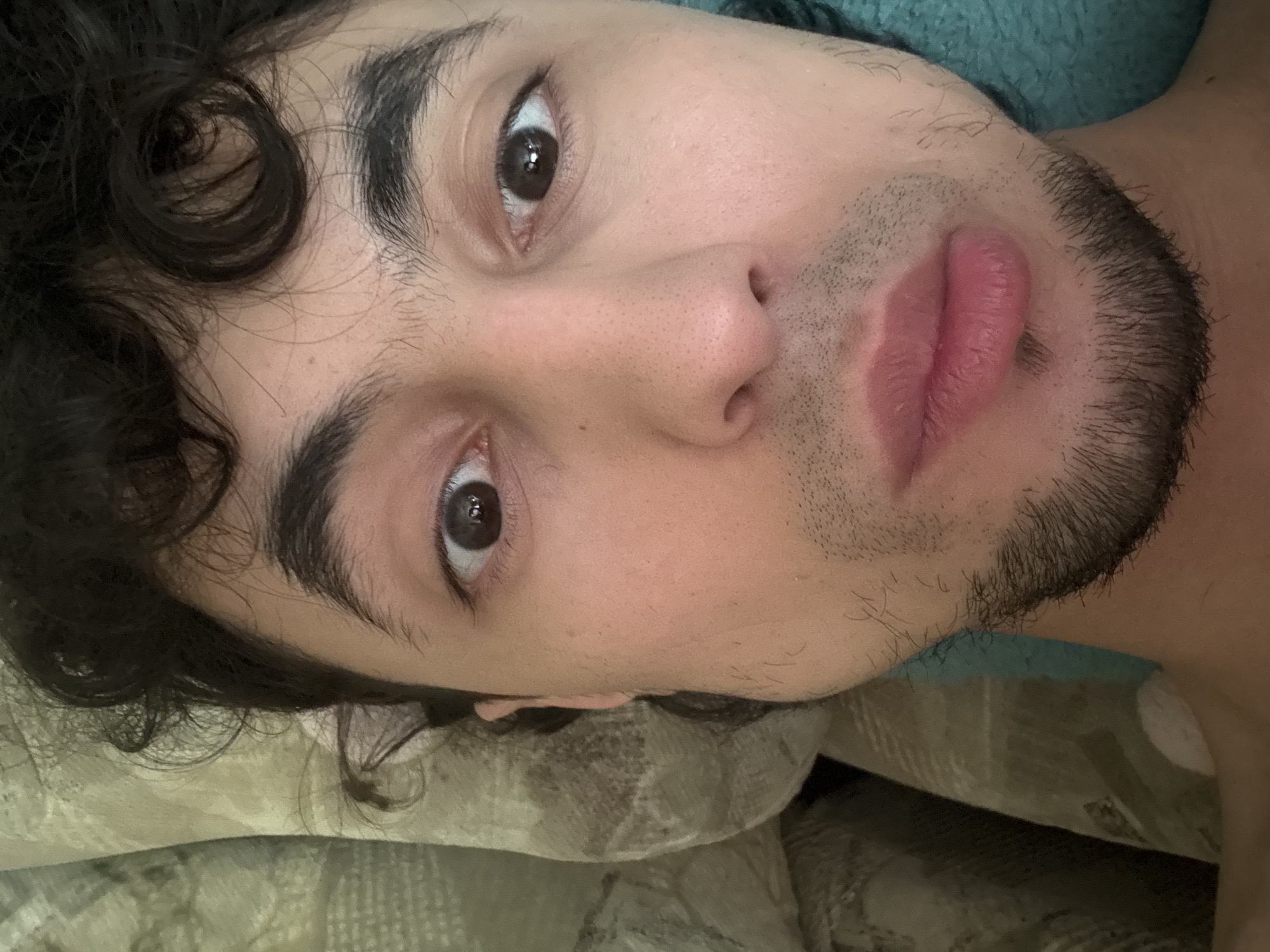 A close-up, Dutch-angle selfie shows a person's face from the eyes down, lying down. The person has dark, curly hair, dark eyebrows, and light skin. Their eyes are open and looking towards the camera. They have a thin mustache and a goatee. Their lips are slightly parted and appear to be a light pink color. The person is lying on a patterned pillow and a teal blanket is visible. The overall lighting is natural and soft.