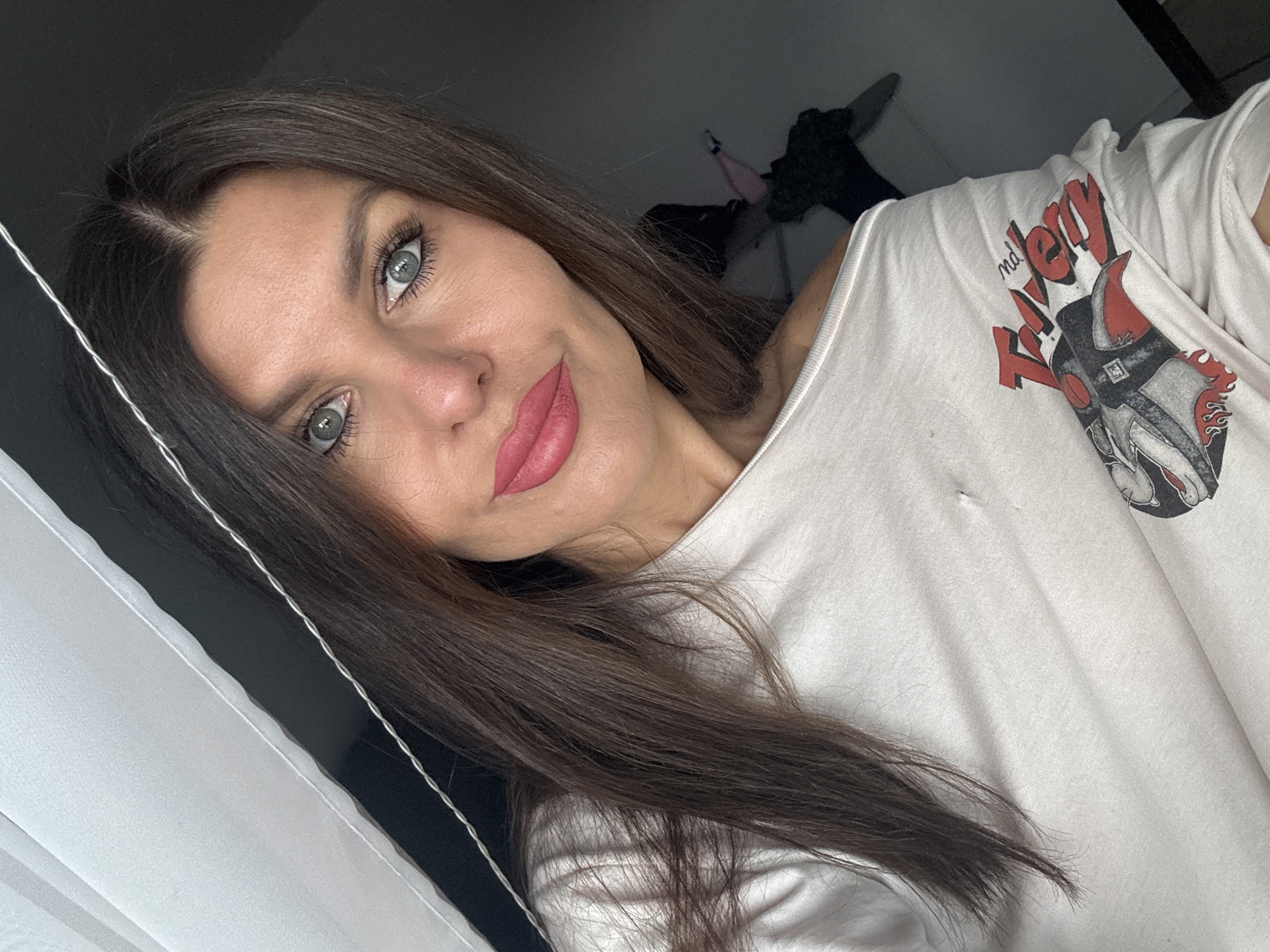 A woman with long brown hair and blue eyes is shown in a close-up shot. She is wearing a white t-shirt with a graphic print on the left side. The t-shirt has an off-the-shoulder neckline. Her makeup includes lipstick and eyeliner. The background appears to be an indoor setting, with a white curtain or sheet on the left and a dark wall on the right. There is a thin, silver wire-like object running diagonally across the top portion of the image. The lighting suggests it is daytime, possibly near a window. The image does not provide any visual cues to determine its location in Chaikovsky, Russia.