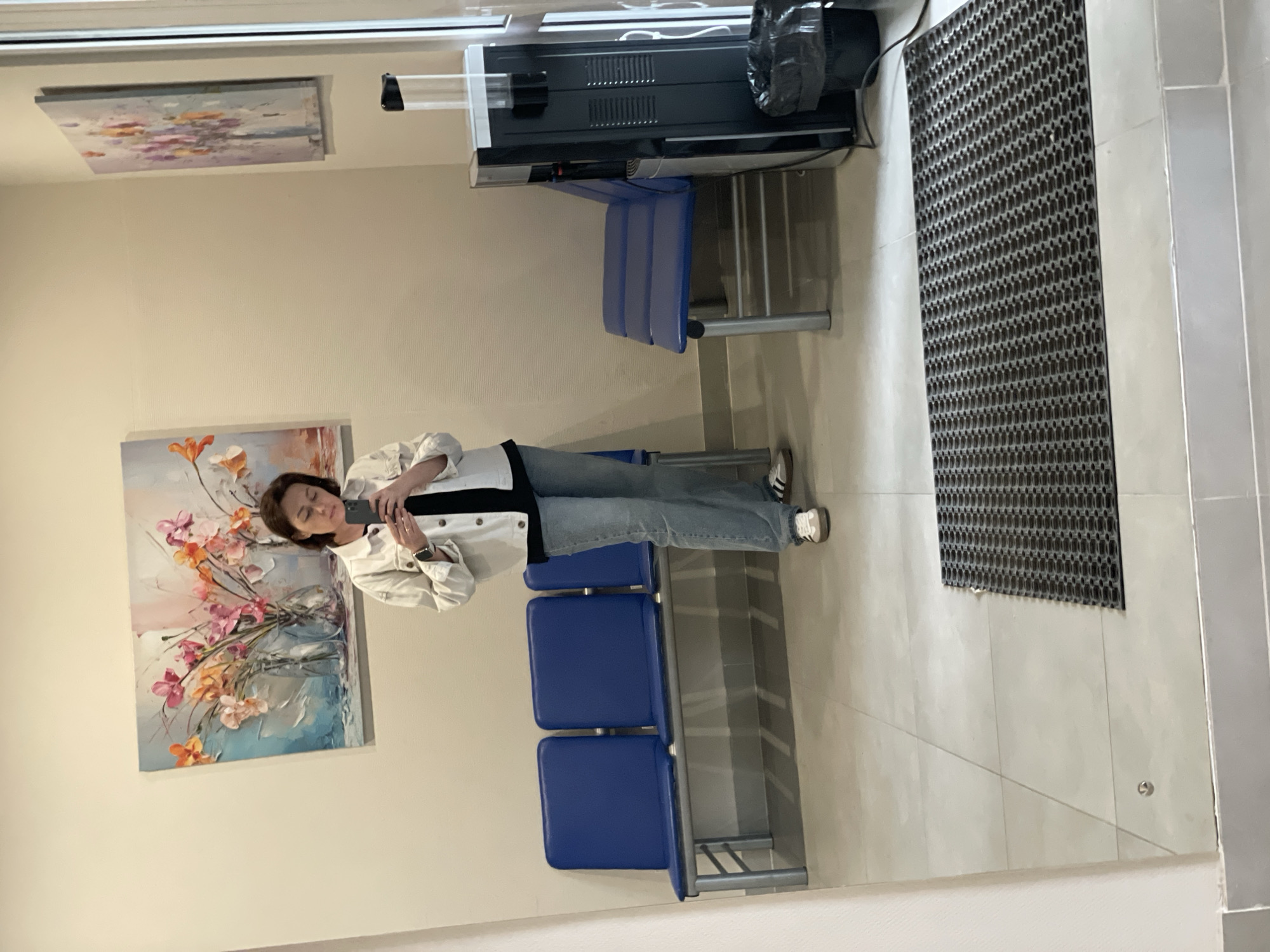 The image, taken in Chaikovsky, Russia, depicts an indoor waiting area or lobby. A woman stands centrally, holding a dark smartphone to eye level, appearing to take a selfie or a photograph of her reflection in a large mirror or reflective surface. She is dressed in a light-colored (possibly white) jacket over a dark top, blue jeans, and white sneakers with dark stripes, and wears a watch on her left wrist.
Behind the woman, against a light-colored wall, are blue public seating benches, each with padded seats and backs on a metal frame. Two rows are visible, one behind the other. On the wall to the woman's left, a framed painting displays abstract floral arrangements in shades of orange, pink, and blue. Higher up and further to her right, another abstract painting features lighter colors, possibly depicting water lilies or a serene landscape.
Above the seating, to the right of the woman, a dark, wall-mounted appliance with vents, resembling an air conditioner or heater, is visible. A black plastic bag is attached to its right side. The floor is covered with light-colored square tiles. A dark, textured rubber mat lies on the tiled floor to the right of the frame. The space appears well-lit, suggesting daytime. There is no visible text in the image. The setting conveys a clean, functional indoor environment.