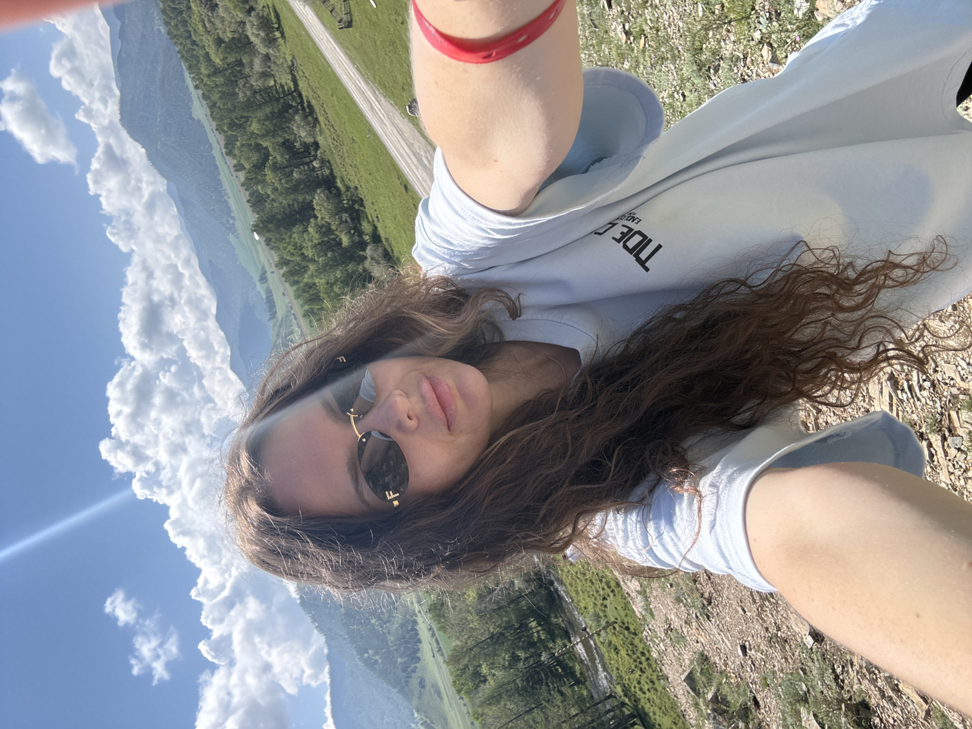 A woman is taking a selfie outdoors in a mountainous, rural area. She has long, curly brown hair and is wearing sunglasses and a light blue t-shirt with a black logo that reads "TIDE OCEAN ENDURANCE". A bright red bracelet is visible on her right wrist. The background features rolling green hills, a dirt road, a body of water, and a range of mountains under a partly cloudy blue sky. The location is identified as Bichiktu-Boom, Russia. The scene appears to be a sunny day.