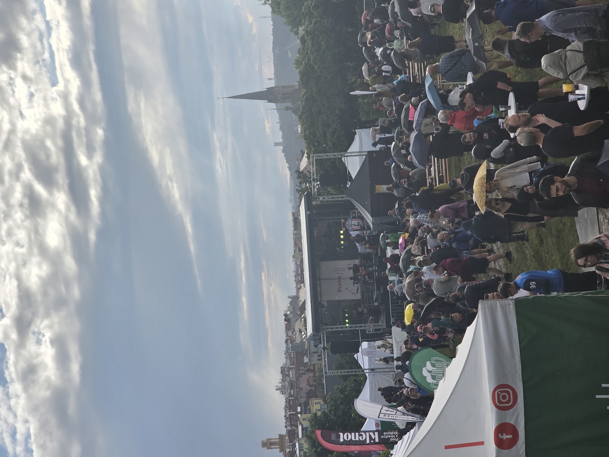 This detailed image captures an outdoor festival or concert taking place under an overcast sky in Prague, Czech Republic. The prominent display of "Praha" on the stage backdrop explicitly identifies the city.
In the immediate foreground on the left, a large white vendor tent with a green lower section and a white upper band is partially visible. Red Facebook ("f") and Instagram logos are printed on its side. The ground throughout the festival area is a grassy field, appearing somewhat worn and possibly damp from rain.
Numerous people, forming a dense crowd, occupy the foreground and mid-ground. Many individuals are holding open umbrellas of various colors, including black, yellow, and blue, indicating either ongoing rain or recent precipitation. Attendees are dressed in casual attire and are seen standing, walking, conversing in groups, and gathered around small, round white tables, some holding drinks in plastic cups. Tiered wooden benches are visible in the mid-ground, providing seating.
A central stage structure, colored black, dominates the mid-ground. A band, comprising at least three musicians with guitars and a drum kit, is actively performing. The stage's backdrop prominently features the word "Praha." To the left of the stage, a tall, red and black vertical banner from a vendor tent is visible, bearing the text "Klenot" and "Pivo z Hradce Králové" (Beer from Hradec Králové), identifying a specific Czech beer vendor. Several other white tents are scattered around the stage and crowd areas.
The background showcases a hazy cityscape, typical of Prague, with a densely packed collection of buildings and rooftops. A distinctive, tall, and slender Gothic-style church spire, topped with a cross, rises prominently above the city skyline on the right side of the frame, serving as a significant landmark. Hills and further distant structures are also discernible beneath the cloudy sky. The overall scene depicts a lively, crowded outdoor event under diffused, possibly damp, lighting conditions.
