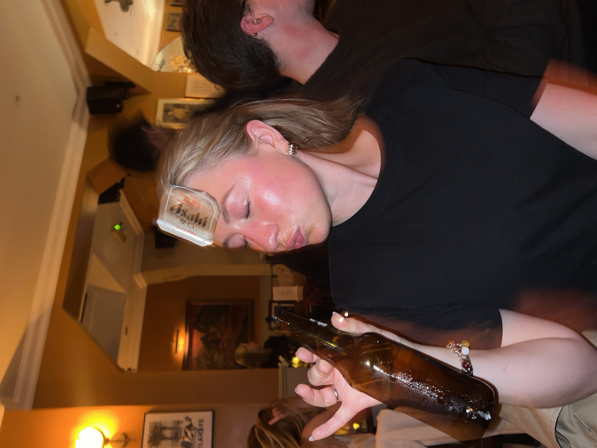 A tilted indoor shot depicts a woman with a beer label adhered to her forehead, holding a brown beer bottle. Her eyes are closed and she's wearing a black top. An individual with dark hair stands behind her. The environment appears to be a bar or pub with muted lighting. Artwork adorns the walls, and portions of furniture are visible. While the image suggests a social setting, specific details about the location in London, United Kingdom, are not discernible from the image.