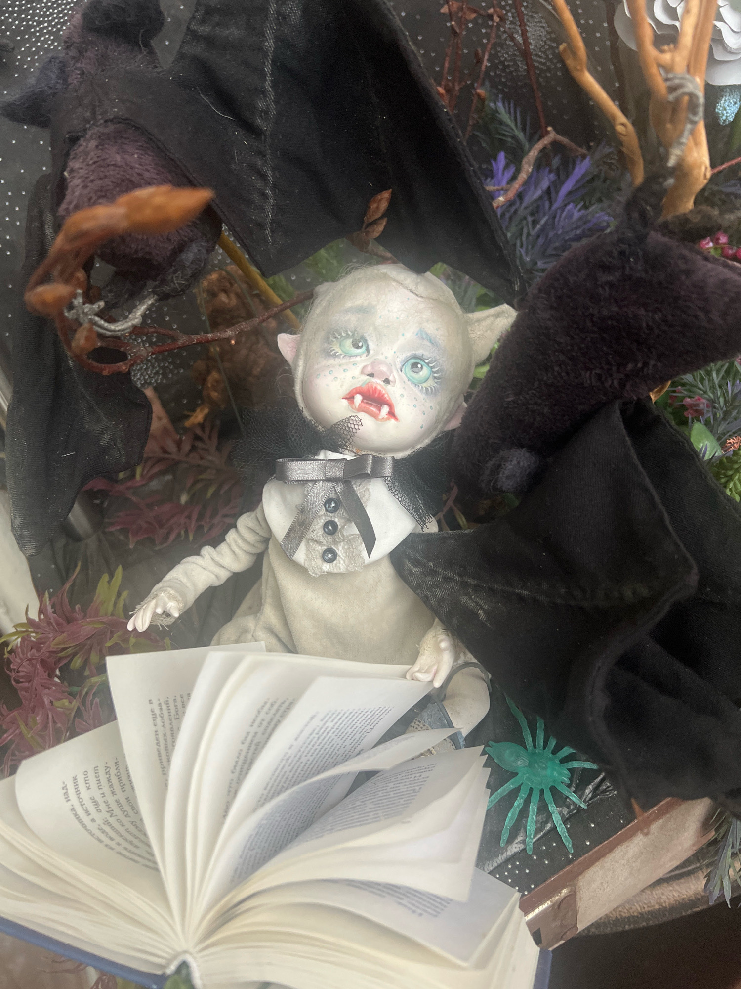 The image depicts a doll that resembles a vampire, with pale skin, pointed ears, and fangs. The doll is wearing a formal outfit consisting of a white shirt, a dark bow tie with buttons, and a light-colored vest. It has large, striking blue eyes and red lips. The doll is seated next to an open book, with its pages fanned out. In the background, there are various Halloween-themed decorations, including dark fabric that resembles bat wings, branches, and artificial foliage in shades of purple and green. A small, green plastic spider is visible on the right side of the image. The scene appears to be a close-up shot of a display, possibly a shop window or a home decoration. There is no discernible information about the location in Zelenogradsk, Russia.