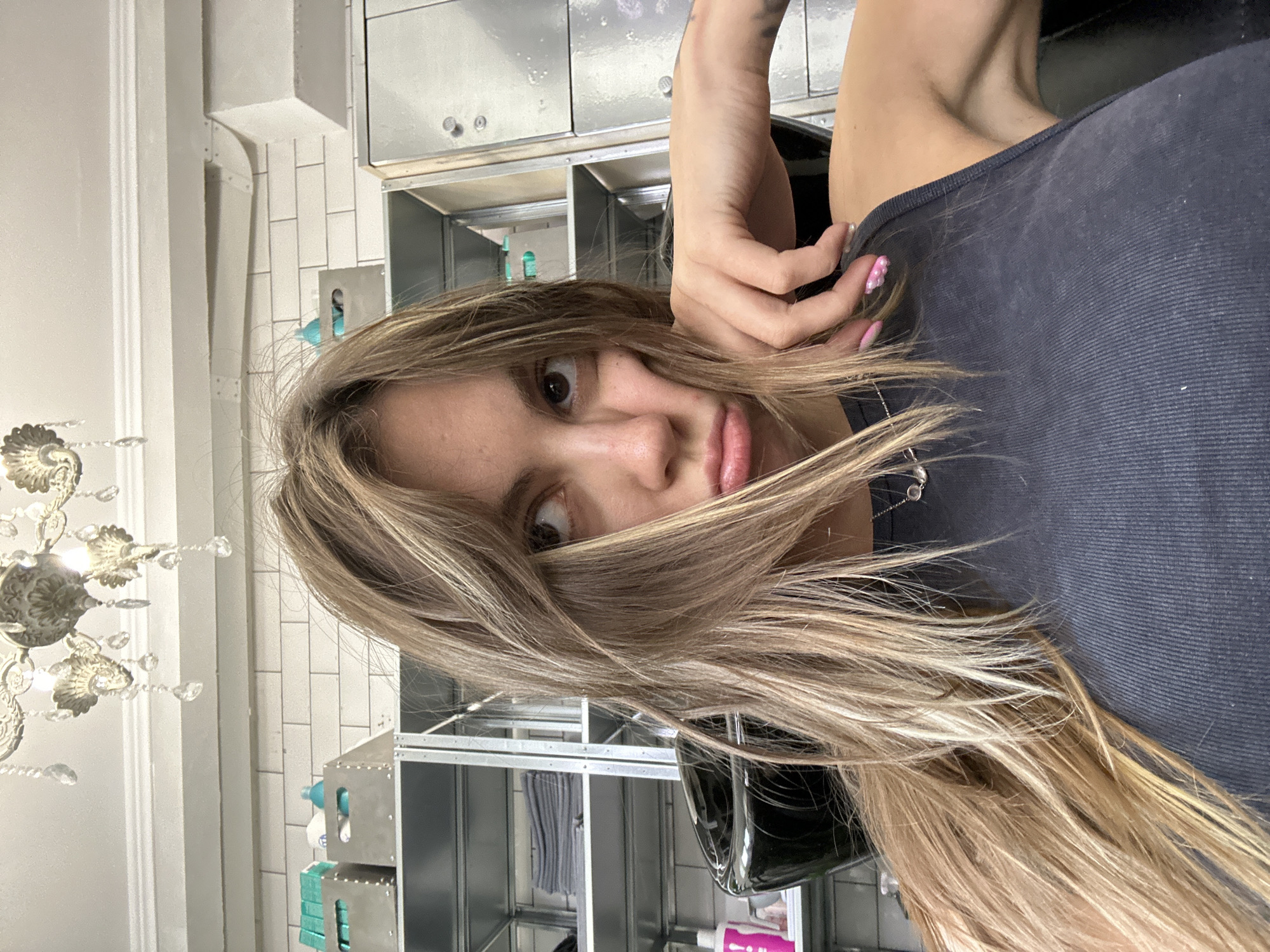 A young woman with long, blonde hair, styled with bangs that partially obscure her face, is pictured indoors. She is wearing a dark grey tank top and a silver necklace. Her face is turned slightly to the side, with her mouth puckered, giving her a pouty expression. Her right hand is resting on her chin, with her fingers touching her cheek. The background appears to be a salon or a similar establishment, with shelves holding various items, possibly hair products or towels. A black basin, characteristic of a hair salon, is visible to her left. Above her head, a chandelier with crystal accents hangs from the ceiling. The walls are tiled with white bricks. The image is a close-up selfie, taken from a slightly high angle. There is no indication of the location being in Moscow, Russia.