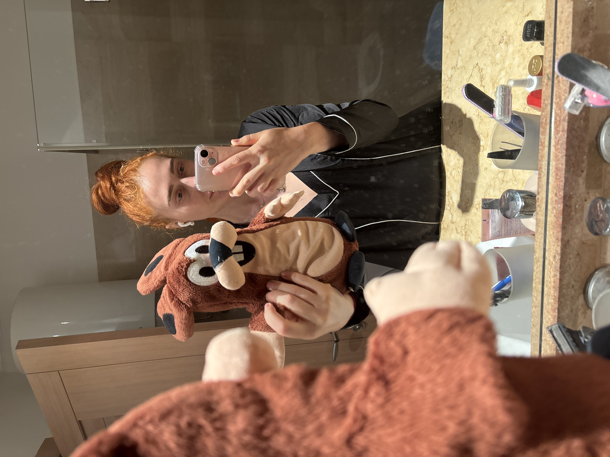 A red-haired woman in black and white pajamas takes a selfie in a bathroom mirror. She is holding a Tazmanian Devil hand puppet, of which a second puppet is also visible in the mirror. The bathroom has a beige marble counter cluttered with cosmetic items and tools. The location is most likely inside a hotel or apartment in Budva, Montenegro.