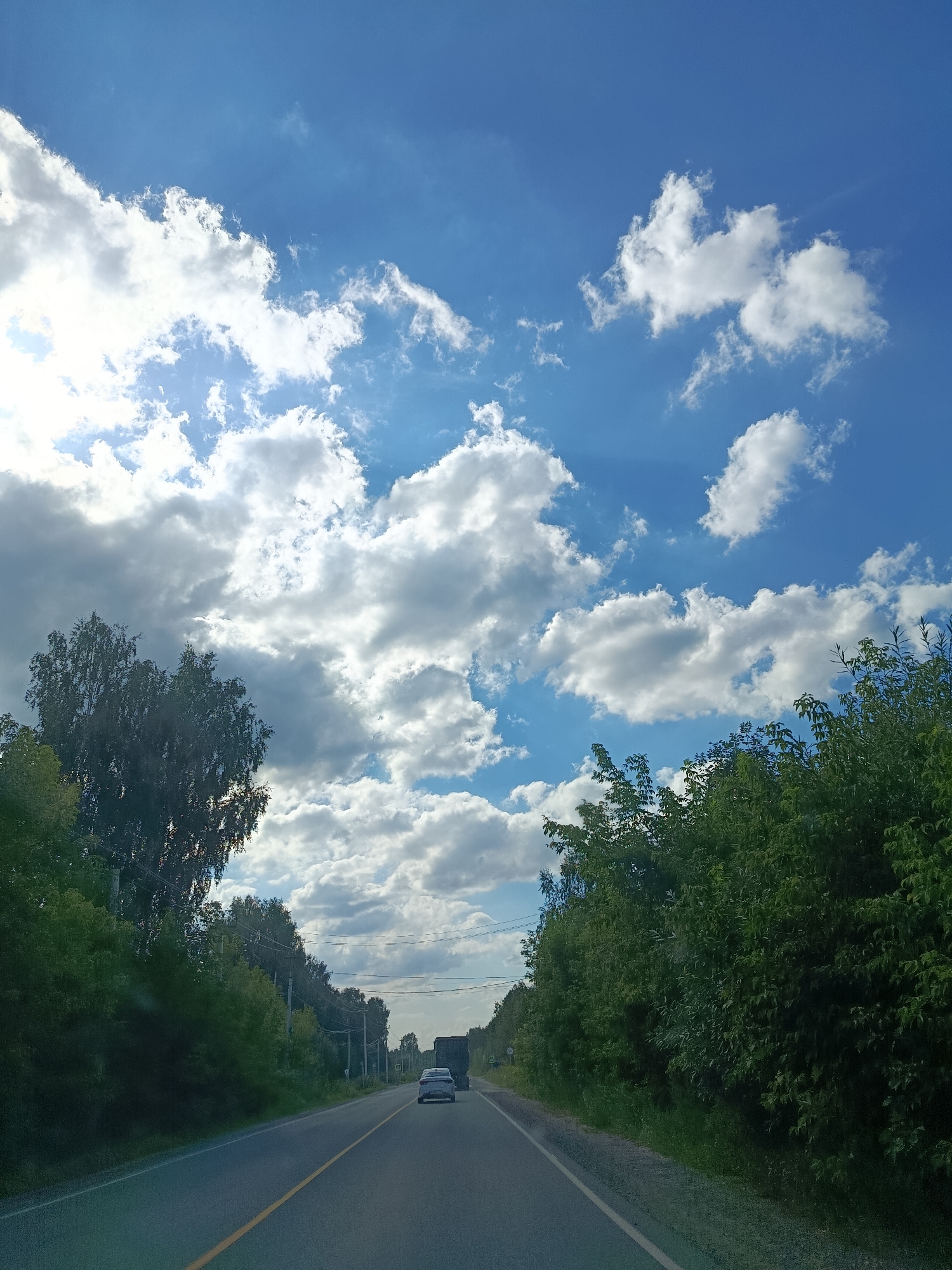 The image shows a two-lane asphalt road stretching into the distance, lined with lush green trees on either side. The road has a yellow line marking the center. Two vehicles are visible on the road: a white car in front and a dark truck behind it. Above the road, a bright blue sky is filled with dynamic white clouds, some of which are illuminated by sunlight. Power lines are visible running along the right side of the road. The scene suggests a daytime view from within a moving vehicle on a rural road in Snopok Novyi, Russia.