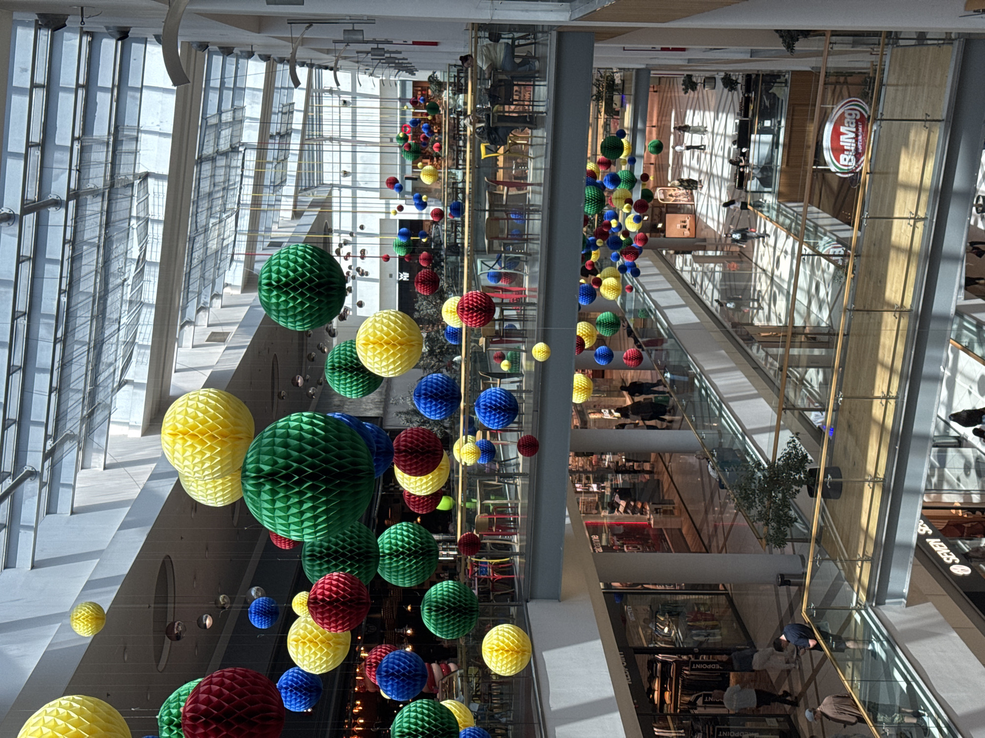 The image depicts the interior of a modern shopping mall in Varna, Bulgaria. The view is from an elevated position, looking down into the central atrium. The architecture is characterized by large glass windows on the left side, allowing ample natural light. Multiple floors are visible, with shops lining the walkways. The walkways are enclosed by glass railings, providing safety and a clear view of the lower levels.
The atrium is decorated with numerous colorful paper lanterns in shades of green, yellow, red, and blue, suspended from the ceiling. These lanterns vary in size and are arranged in a seemingly random pattern, adding a festive and whimsical element to the scene.
Several people can be seen on different levels of the mall, engaging in activities such as shopping and walking. Some people are standing near shop displays, while others are walking along the walkways. A "BulMag" sign is visible on one of the storefronts.