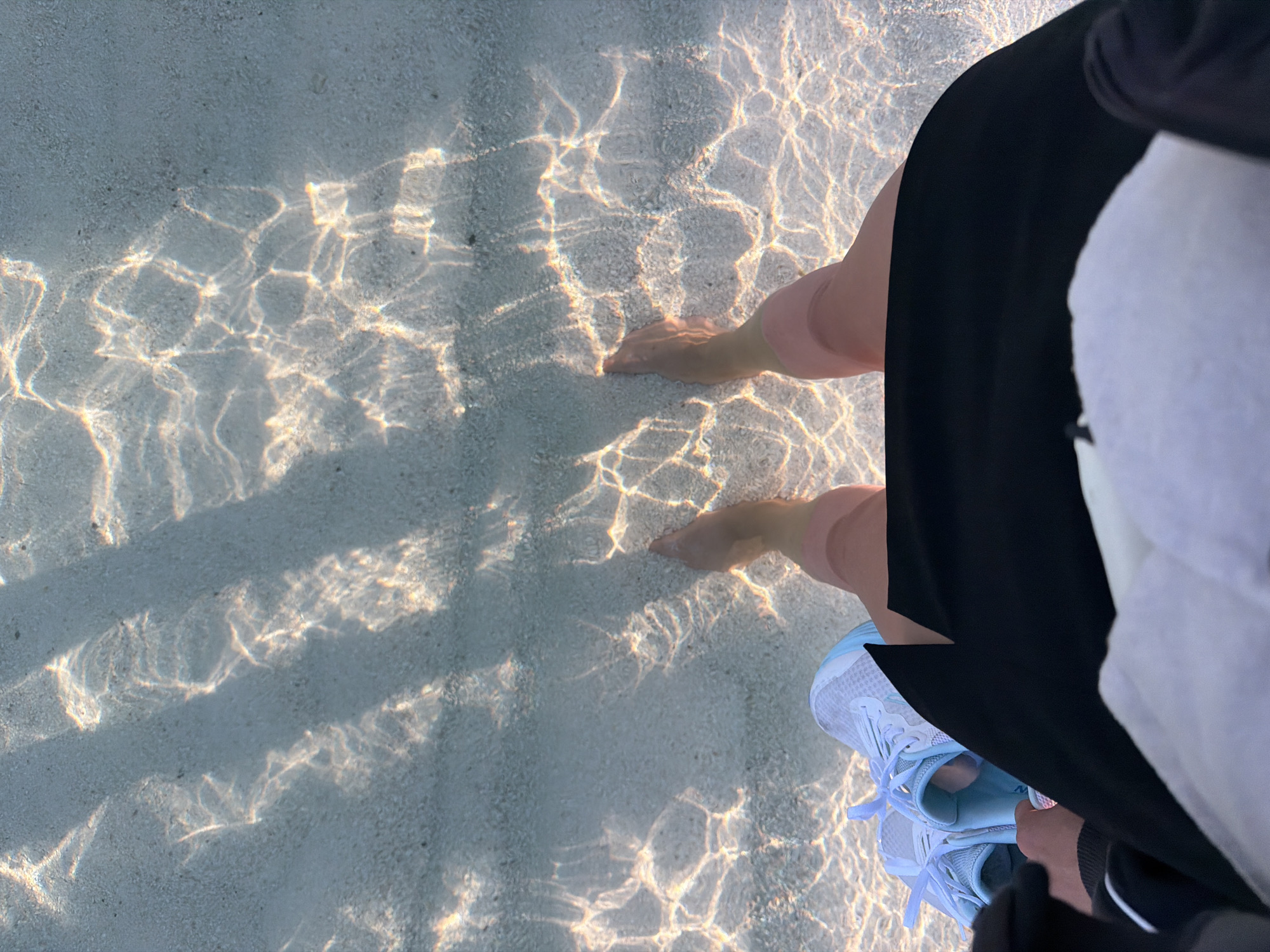 A first-person perspective shot shows a person's legs and feet submerged in clear shallow water, likely at a beach in Ksamil, Albania. The sunlight refracts through the water, creating rippling patterns on the sandy seabed. The person is wearing a black skirt and white socks. A pair of light blue athletic shoes are placed to the left of their feet, near the edge of the water. The background is obscured by the shallow water and the person's clothing.