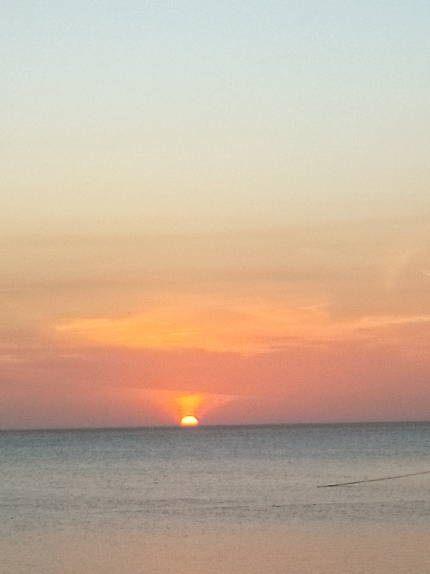 This media file captures a tranquil sunset scene over a vast body of water, likely the ocean, in Juan Griego, Venezuela. The horizon line cleanly divides the sky from the sea, creating a balanced composition.
The sky is a prominent feature, displaying a beautiful gradient of colors characteristic of sunset. At the very top, the sky is a soft, pale blue, transitioning smoothly into a warm, light yellow-orange in the mid-section. Closer to the horizon, the colors intensify to vibrant oranges and deep reds, with subtle streaks of clouds diffusing the light. The sun itself is partially visible, appearing as a bright, glowing orange-yellow arc just above the horizon line, casting a radiant glow.
Below the horizon, the sea stretches out, appearing calm and still. Its surface reflects some of the sky's subdued light, exhibiting muted tones of blue-gray with hints of the warm oranges from the sunset closer to the horizon. There are no distinct waves visible, contributing to the serene atmosphere. In the lower right portion of the water, a faint, dark, horizontal line can be observed, which might be a distant object, a reflection, or a wake.
The overall impression is one of peaceful natural beauty at dusk. There are no people or discernible man-made structures present, emphasizing the untouched quality of the moment. No text is visible in the image.
