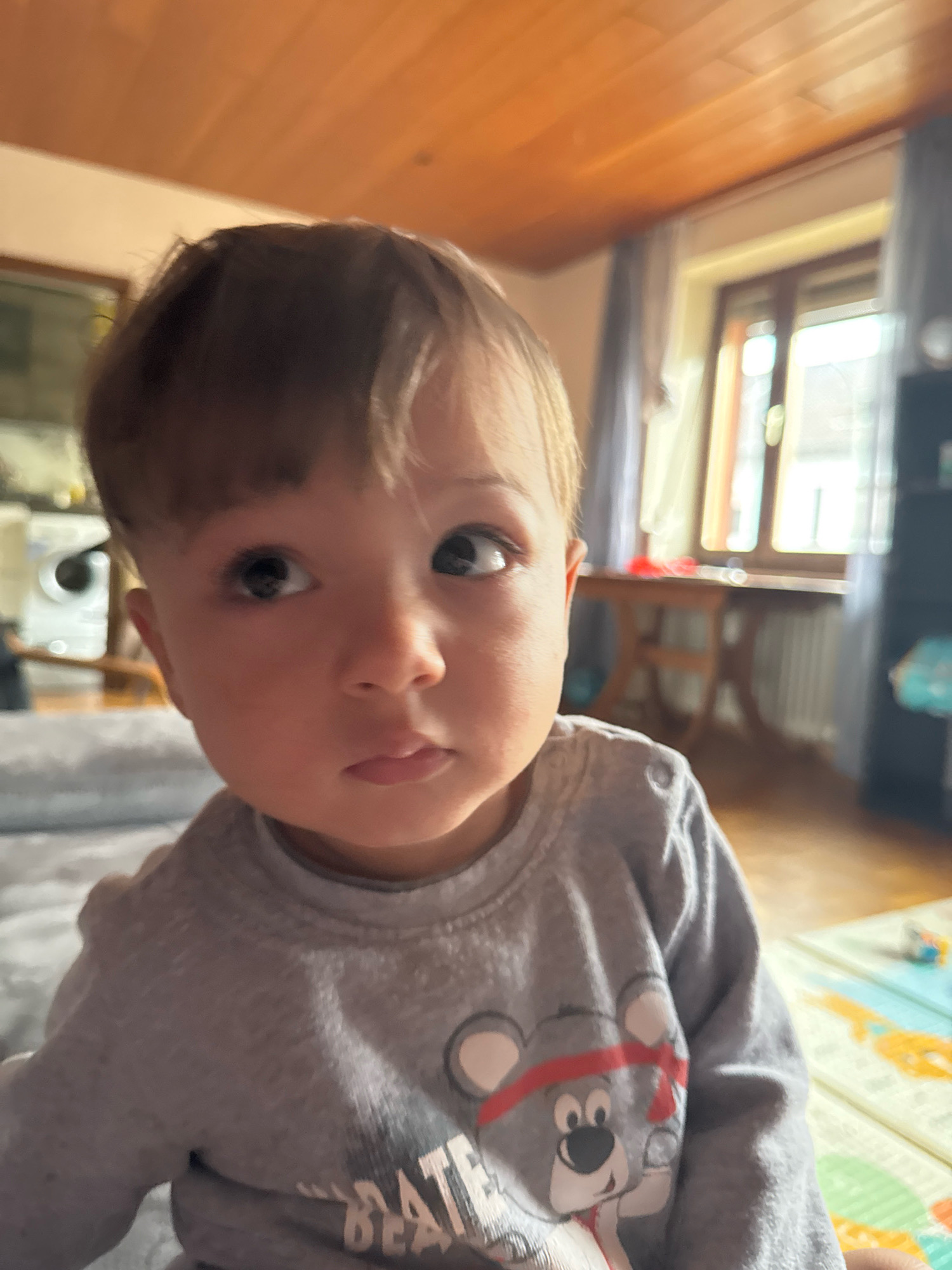 A close-up, waist-up shot shows a light-skinned baby looking up and to the right, with their head tilted slightly. The baby has light brown hair and wide, dark eyes. They are wearing a grey long-sleeved shirt with a cartoon bear graphic. The background is softly blurred, showing a room with wooden ceiling panels, a window with a wooden frame, and a table and chair. A mat with colorful geometric patterns is visible on the floor in the lower right corner. The scene appears to be indoors, likely a residential setting. The location is identified as Überherrn, Germany.