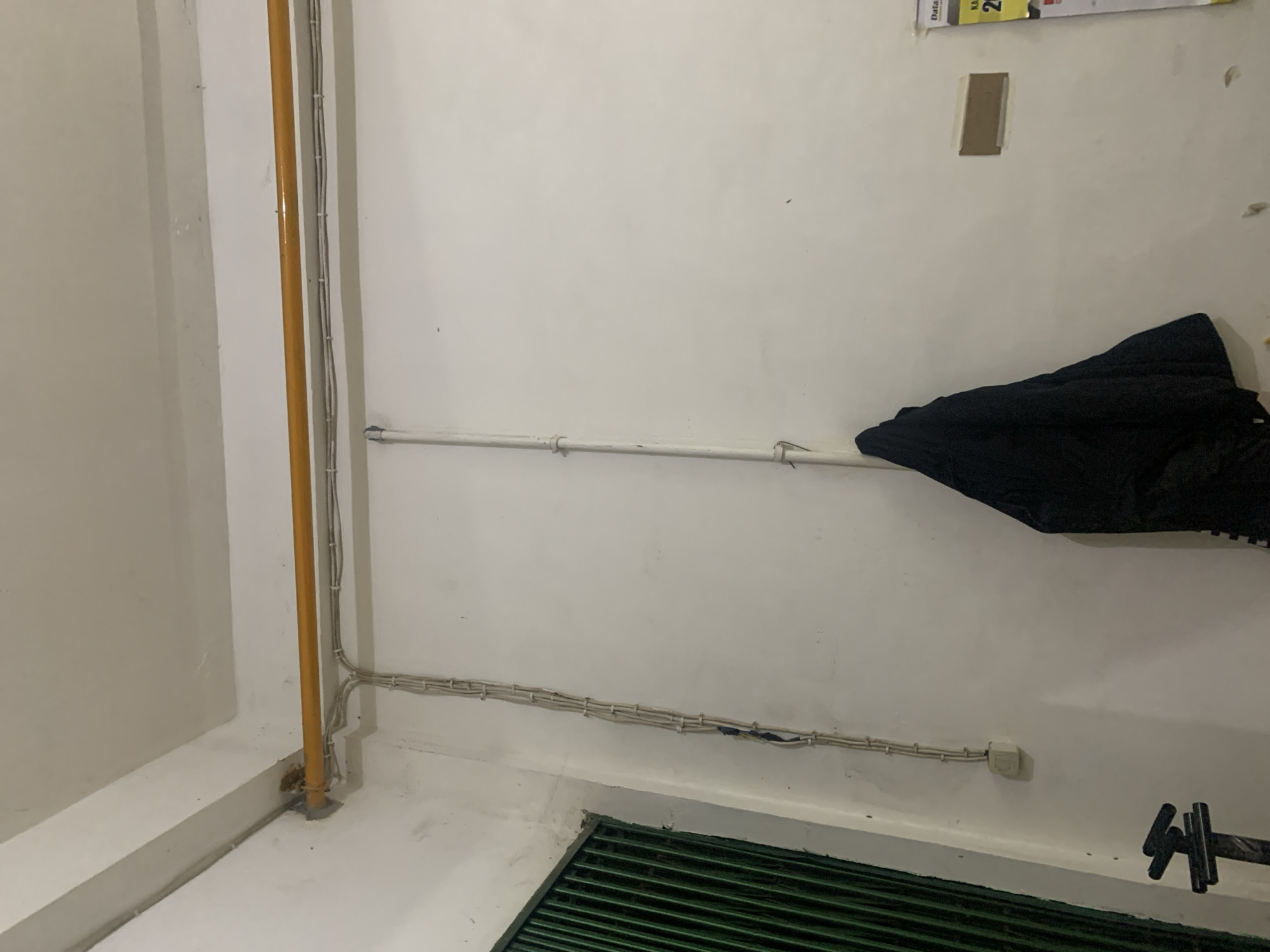 The image depicts an interior scene with a white wall and ceiling. A yellow pipe runs horizontally along the top of the wall, connected to a conduit carrying electrical wires that descend vertically. To the left, a dark green metal gate or shutter is partially visible. A black jacket is hanging on a white vertical pipe on the wall. A calendar with Indonesian text and the number "29" is attached to the wall on the right. The overall impression is of a utilitarian or industrial indoor space, possibly a storage area or workshop, within a building in Palembang, Indonesia.